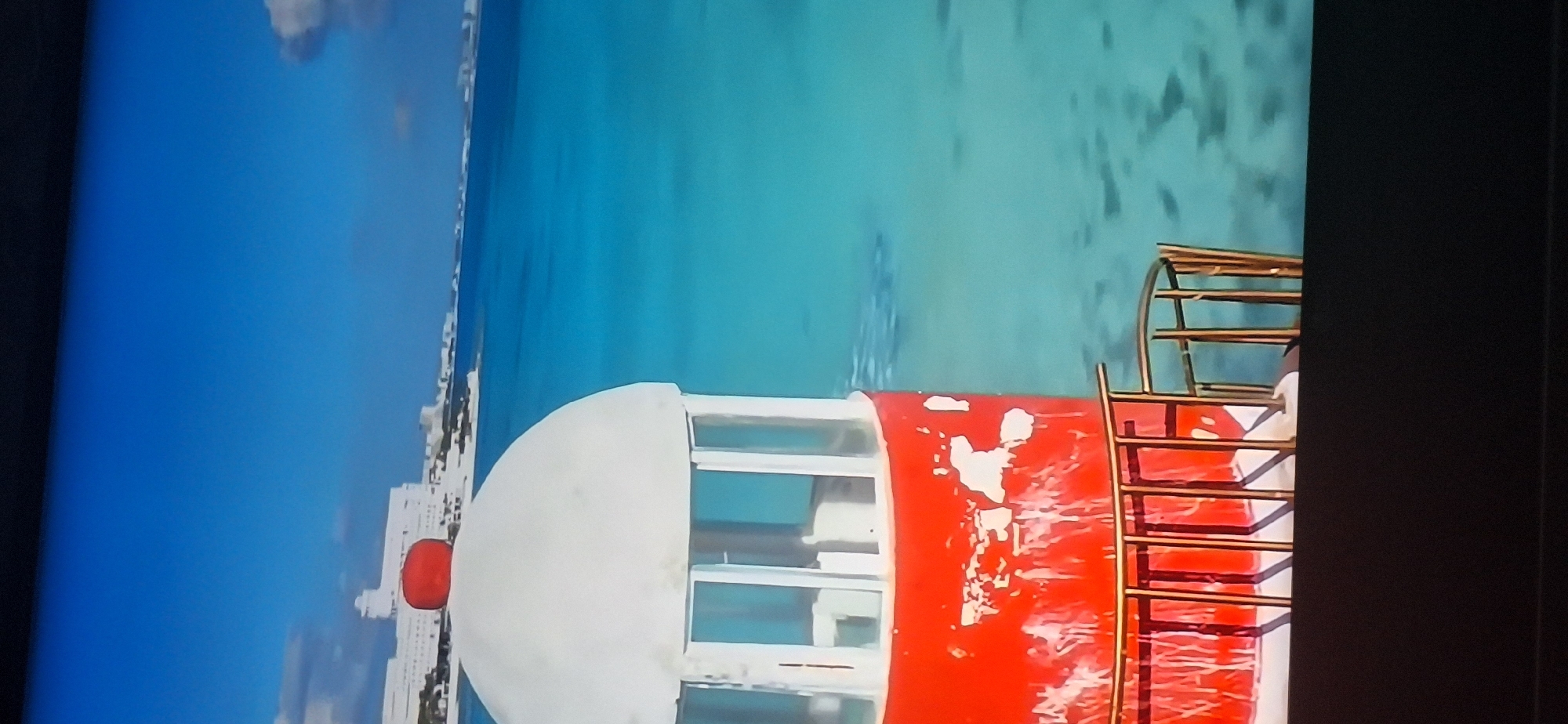 A tall, red and white lighthouse stands on the right side of the frame, its red base peeling in places. A railing surrounds the base of the lighthouse. The lighthouse is positioned in front of a bright blue, clear ocean. In the distance, a white city skyline is visible. The sky is a clear, vibrant blue. There are no people or animals visible in the image. The scene is likely a coastal resort or city. This image is not in Bila Tserkva, Ukraine, as Bila Tserkva is a landlocked city.