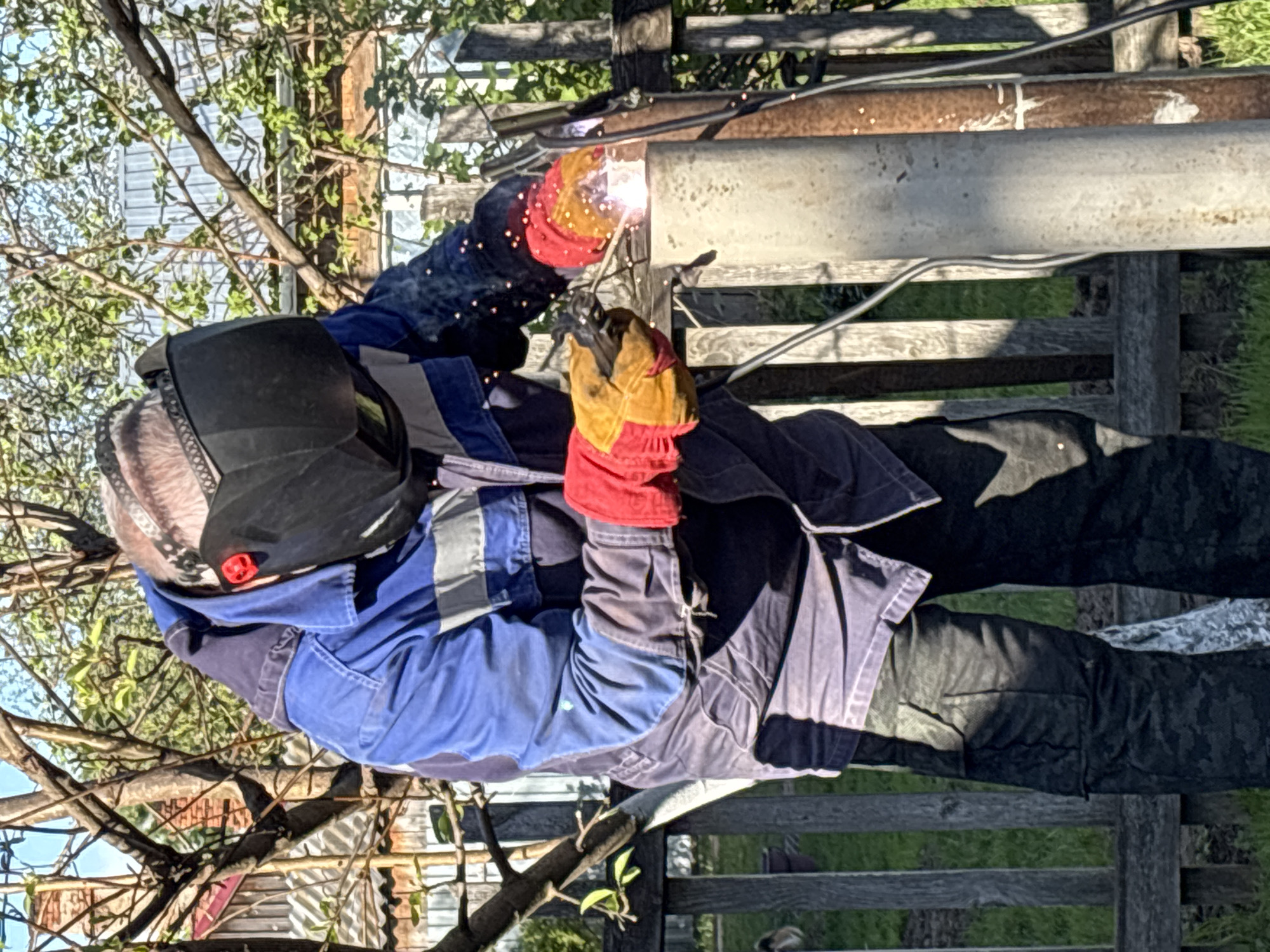 A man is welding a pipe in an outdoor setting in Ваньки, Russia. He is wearing a welding mask, red and yellow gloves, and a blue jacket, and dark pants. Sparks are visible from the welding activity. The welding is taking place on a rusty pipe supported by a wooden fence. There are trees and vegetation in the background, and what appears to be a building with light-colored siding is also visible.