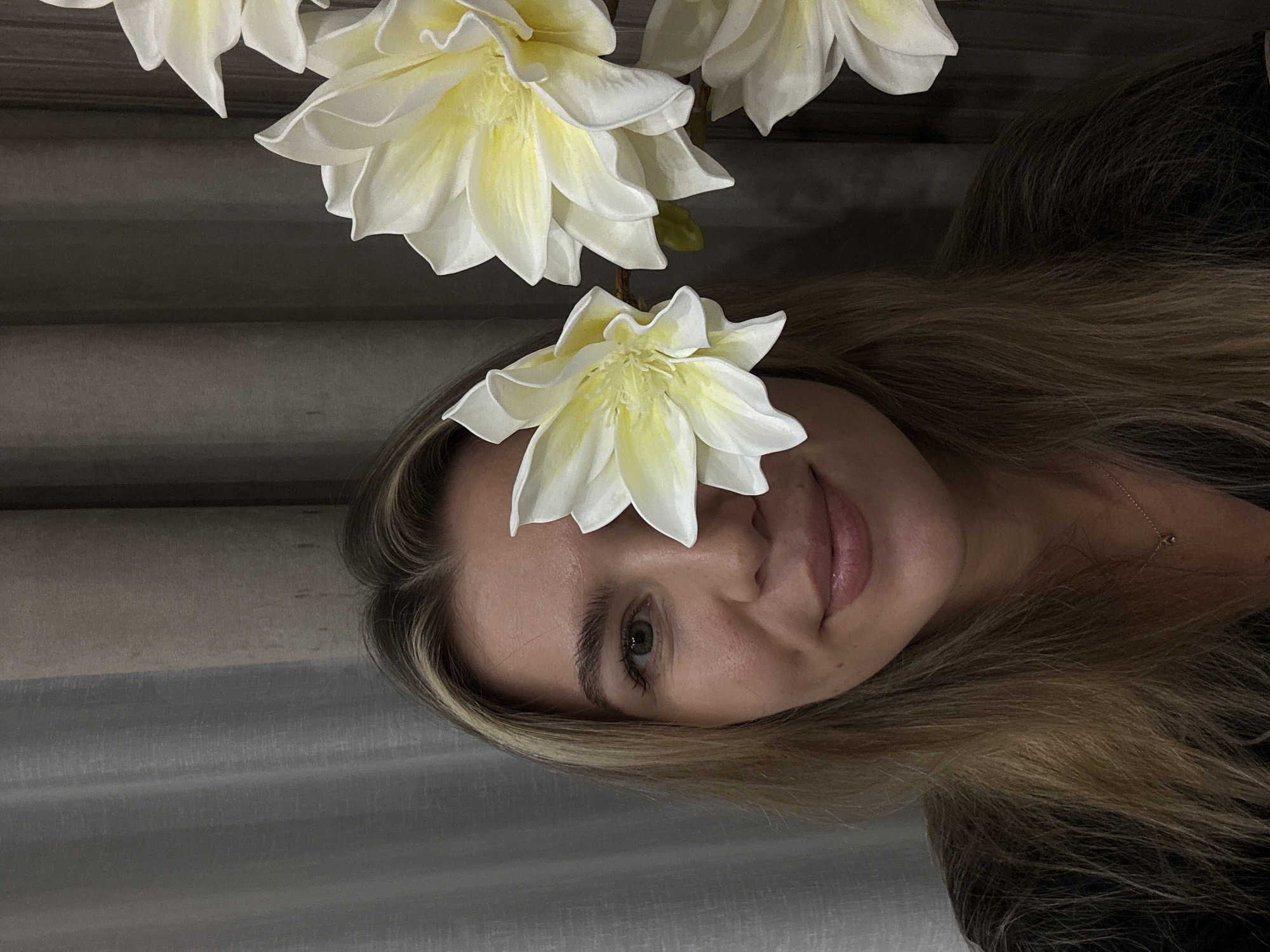 A young woman with blonde hair and light skin is shown from the chest up in a portrait. She is smiling, and a white flower is held in front of her right eye, obscuring it. She wears a dark top and a delicate necklace. The background consists of a textured curtain and a dark wall. Artificial white and yellow flowers are visible on the right side of the image. The lighting is dim.
