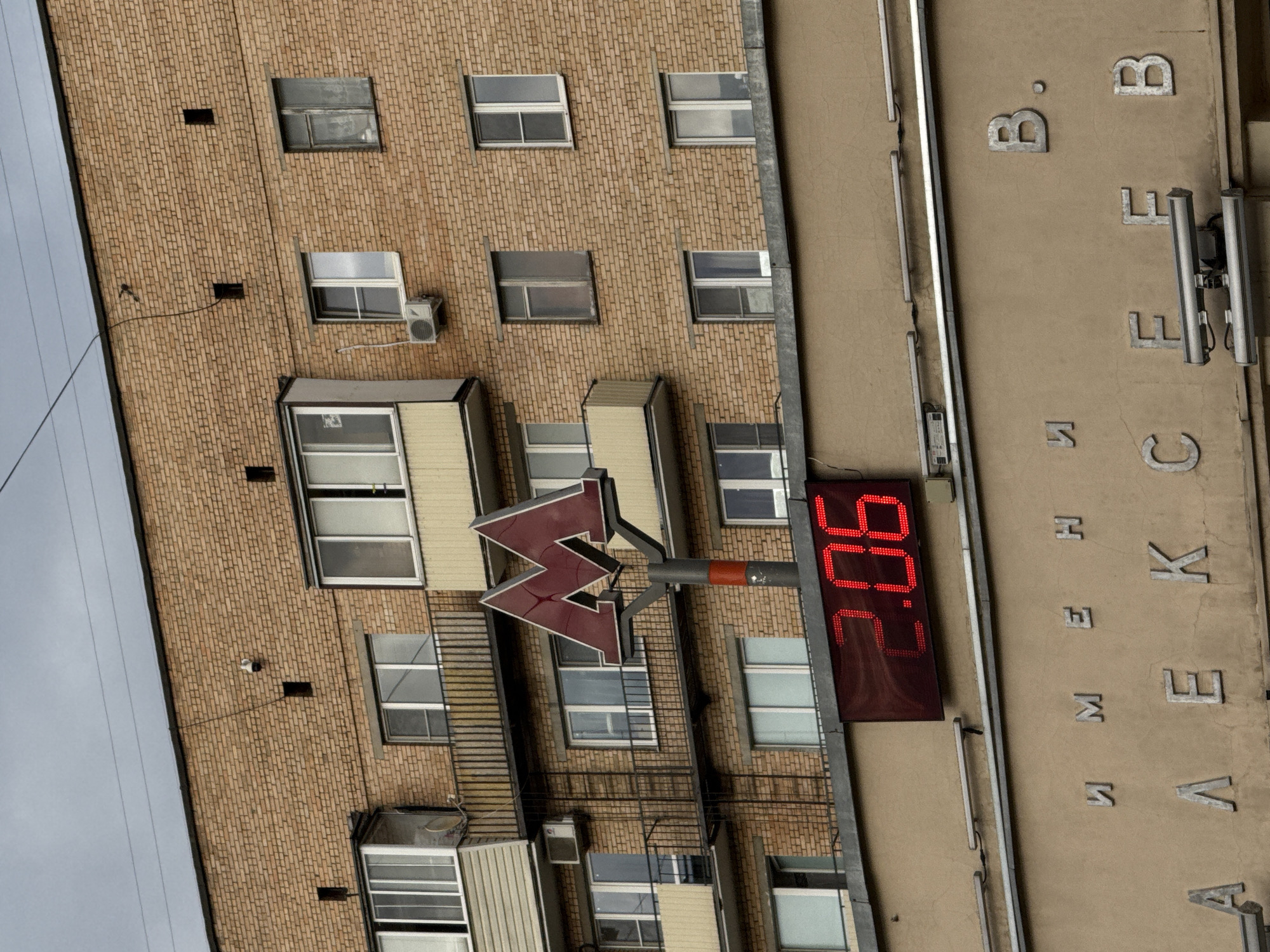 The image depicts the exterior of a building in Moscow, Russia, captured from a tilted perspective. The main subject is a multi-story brick building with a facade featuring numerous rectangular windows; some are partially open, while others reflect the overcast sky. Several white air conditioning units are mounted on the brick wall below some of the windows.
Prominently displayed on a pole extending from the brick building is a large, red, illuminated "M" symbol with an upward-pointing arrow, characteristic of a Moscow Metro (subway) station sign. Adjacent to the Metro sign, a red digital LED display shows the number "90.2".
To the right of the brick building, another structure or wall, light brown in color, is visible. Affixed to this wall are several individual silver-colored Cyrillic letters. These include "B", "B.", "E", "K", "C", "A", "M", "И", and "Л". The letters are arranged in a somewhat scattered vertical fashion, and due to the image's tilt, some appear upside down relative to a normal reading orientation, suggesting they are part of a larger, partially visible sign.
The sky on the left side of the image is grey and overcast, indicating a cloudy daytime scene. No people or specific activities are visible, presenting a static urban architectural view.