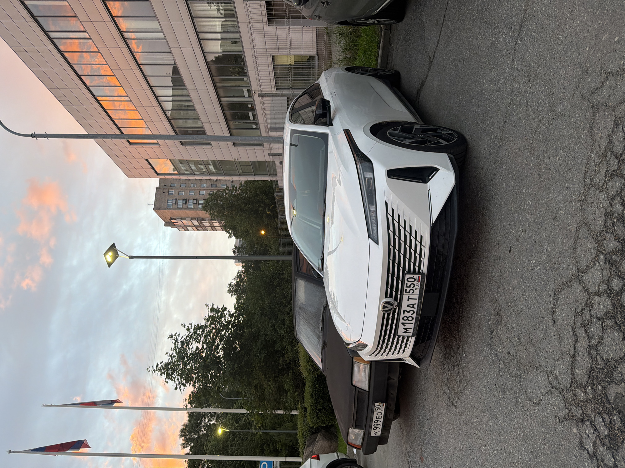 The image shows a white sedan parked in a lot, with a darker colored sedan parked behind it. Both cars have license plates visible. The white sedan is in the foreground, and its front end, including the grille and headlights, is clearly visible. The darker sedan is partially obscured by the white sedan. In the background, there is a modern building with large windows that reflect the sky. Two Russian flags are flying on flagpoles to the left of the frame. The sky has some clouds, and the lighting suggests it is either early morning or late afternoon. The ground appears to be asphalt, with some cracks. There are trees and bushes visible around the parking lot and between the cars.