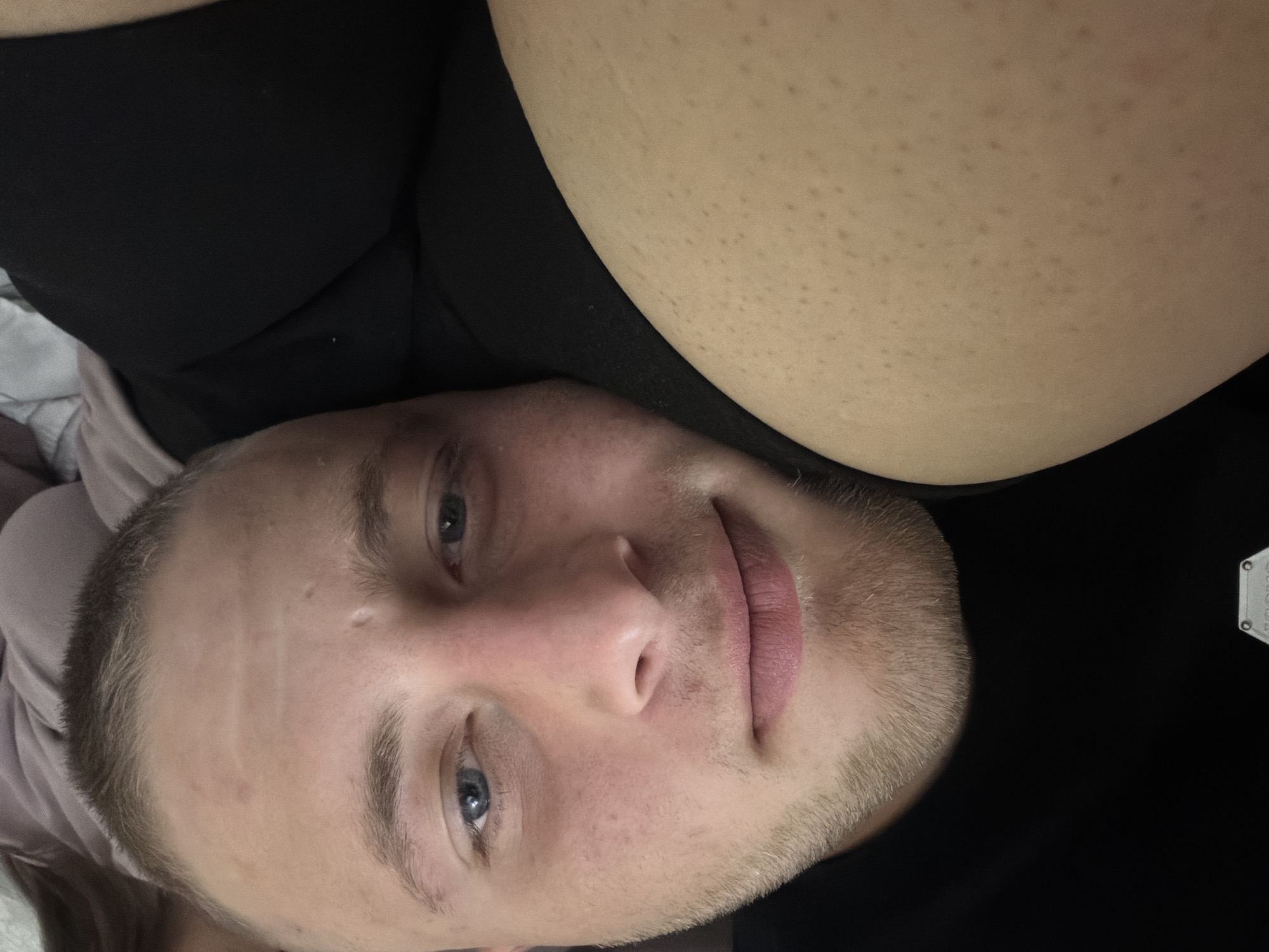 A close-up, over-the-shoulder, selfie-style shot shows the face of a young Caucasian male with light skin and blue eyes. He has short, light brown hair and a light stubble beard. His head is resting on a person's thigh, which is visible in the foreground and has many small, dark freckles or pores. The man is looking directly at the camera with a slight, relaxed smile. He is wearing a black t-shirt. The background is dark and out of focus, with hints of a light-colored garment to the right, suggesting an indoor setting. The image's perspective and focus on the individual's face and upper body indicate a personal, candid moment. No specific elements place the image within the city of Strășeni, Moldova.