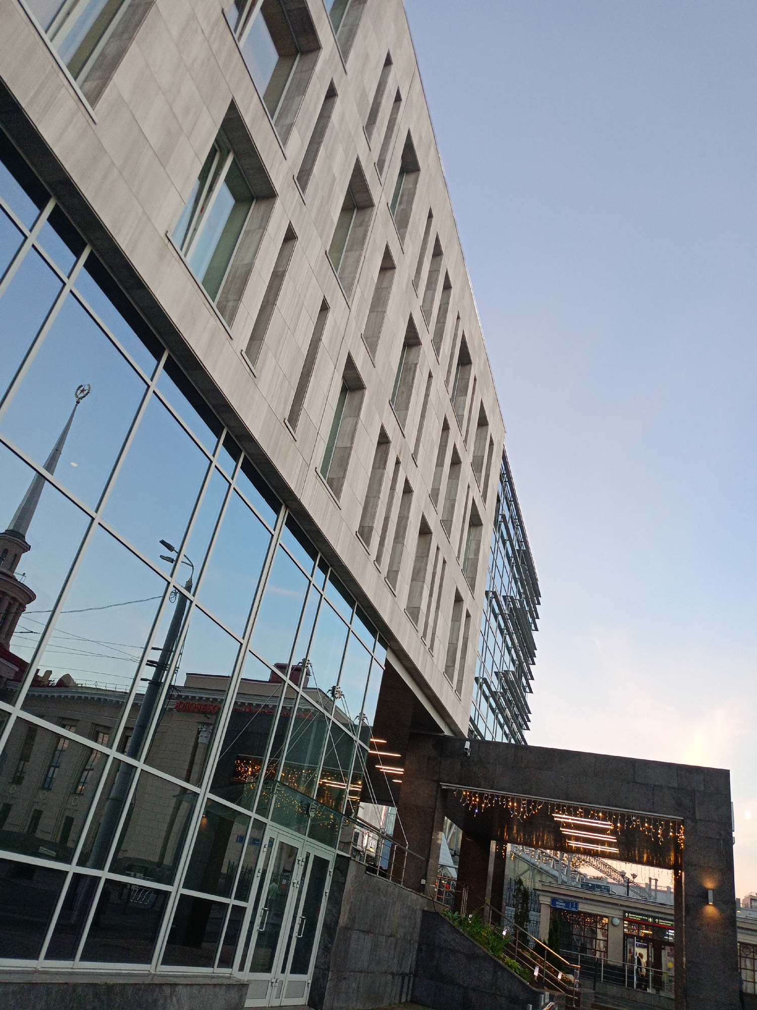 This is a low-angle, wide shot of a modern building with a reflective glass facade and a grey stone exterior. The building appears to be in an urban setting, as indicated by the reflection of an older building with a spire and a star on top in the glass. The sky is a clear, pale blue, suggesting it is either early morning or late afternoon. The facade has numerous vertical, narrow windows. At the base of the building, an entrance is covered by a dark stone overhang decorated with string lights. A set of stairs with metal railings leads up to this entrance. The reflection in the glass also shows a street, a lamppost, and a glimpse of shops or businesses. The overall impression is one of contemporary architecture contrasting with older structures.