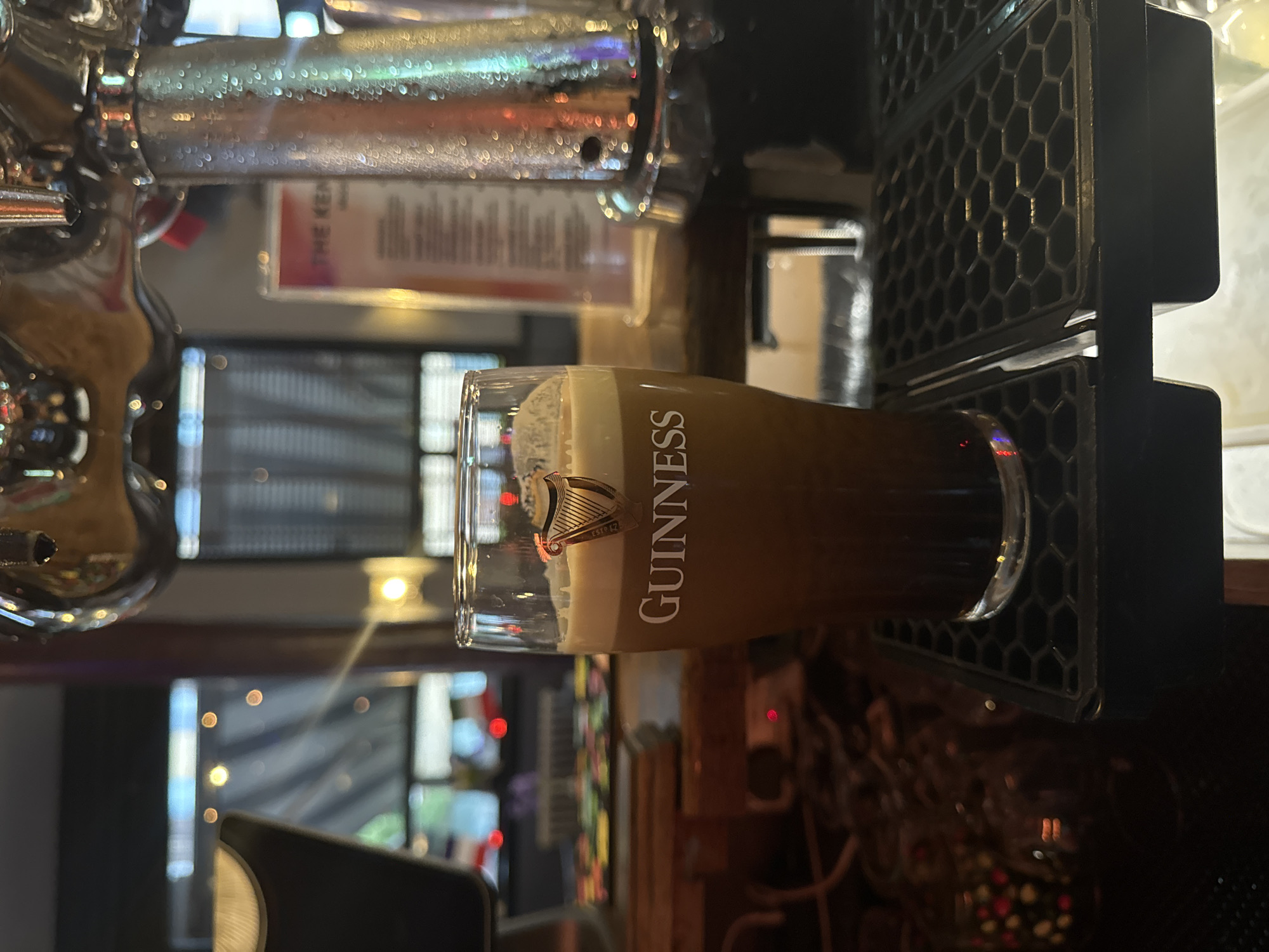 A glass of Guinness sits on a bar top in a pub. The Guinness tap is visible in the background. The pub's interior is blurred but windows are visible. The city is not identifiable from this image.