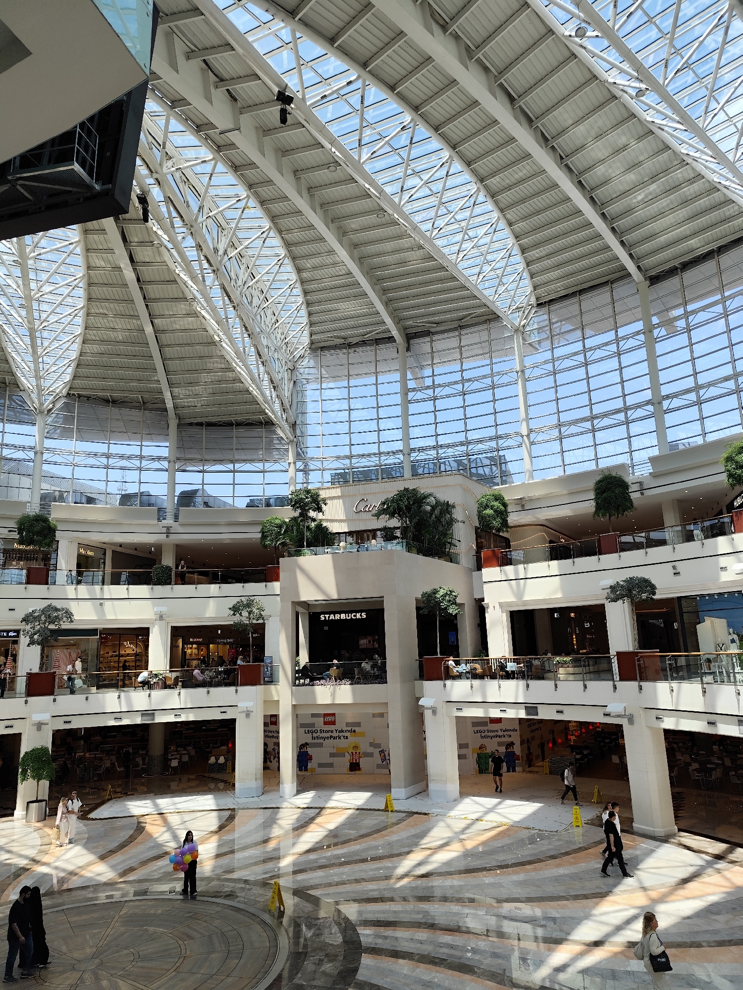 The image depicts the interior of the İstinyePark shopping mall located in Sarıyer, Turkey. The mall features a large, multi-story open space covered by a high, arched glass and metal roof, allowing ample natural light. The interior architecture includes multiple levels of retail spaces, connected by walkways and open areas. A prominent Starbucks is visible in the central area, along with other recognizable store names such as Cartier, Max Mara, and LEGO. A LEGO store under construction is indicated by a sign stating "LEGO Store Yakında İstinyePark'ta", which translates to "LEGO Store Coming Soon to İstinyePark". People are present throughout the mall, some walking, shopping, or seated at outdoor cafe areas. In the foreground, some individuals are gathered near a fountain or decorative area. The floor is patterned with circular designs. Yellow warning cones are scattered on the floor in the vicinity of the LEGO store. The overall scene is a bustling and modern shopping environment.