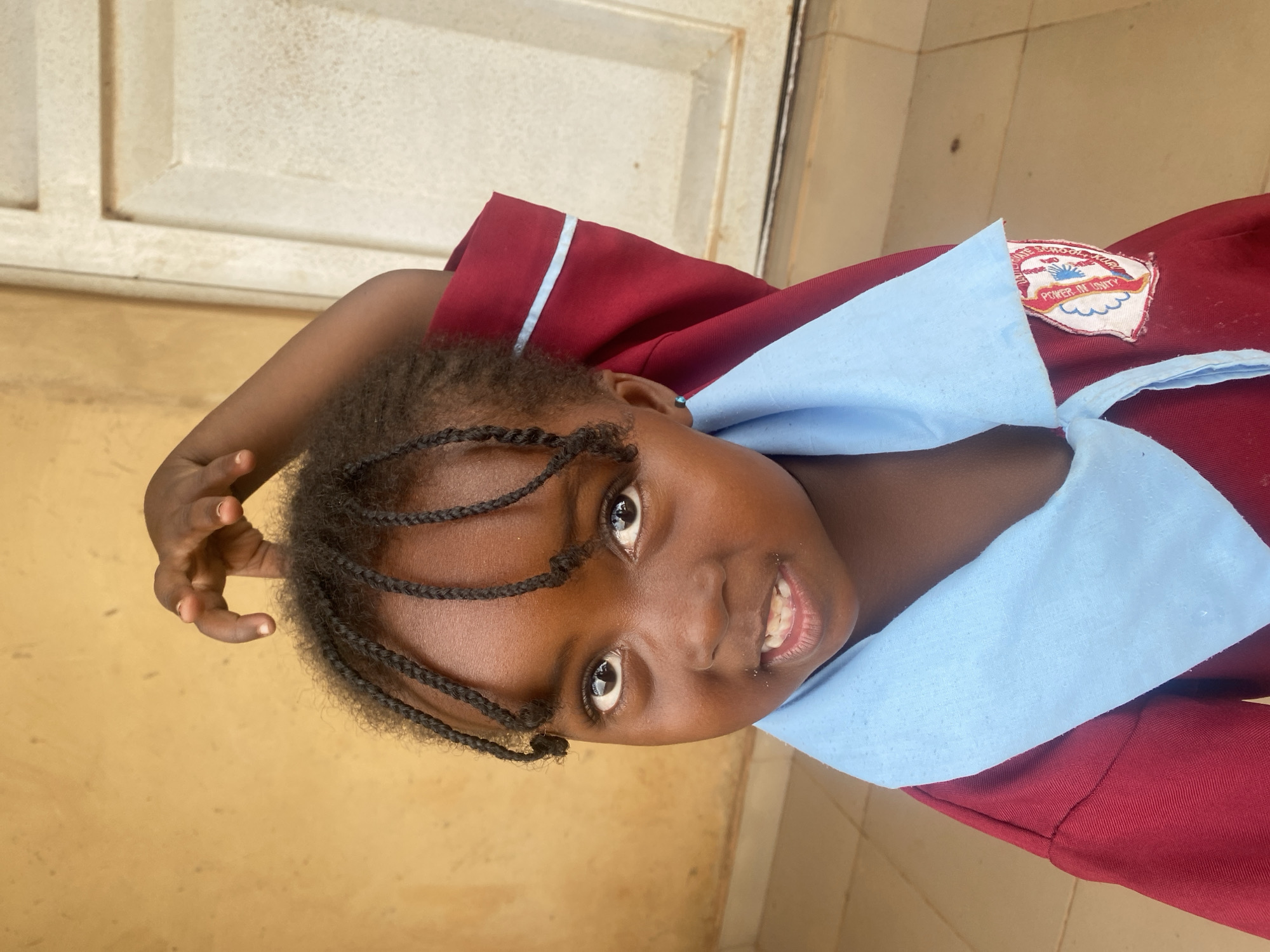 A close-up, portrait-style photograph captures a young girl, likely of African descent, smiling brightly and looking directly at the camera. She has dark skin and her hair is styled in numerous small, dark braids that frame her face. Her eyes are dark and engaging, and a slight, open-mouthed smile reveals her teeth.
She is wearing a school uniform, which is primarily maroon with a light blue collar and a light blue stripe visible on the edge of the sleeve. On the left side of her chest, a school emblem is prominently displayed. The emblem is white with red and blue elements, and the text "POWER IN UNITY" is clearly visible within a red banner. Partially visible above this text is "SCHOOL..." suggesting part of the school's name.
The girl's right arm is bent, with her hand raised near her head or shoulder, possibly in a playful gesture. The background consists of a plain, light brown or tan wall or floor, with a section of a lighter, off-white panel or door visible in the upper part of the image, suggesting an indoor or covered outdoor setting. The lighting is soft and even, indicating daytime. The scene conveys a sense of youthful cheerfulness. The location is Kuru Dungus, Nigeria.