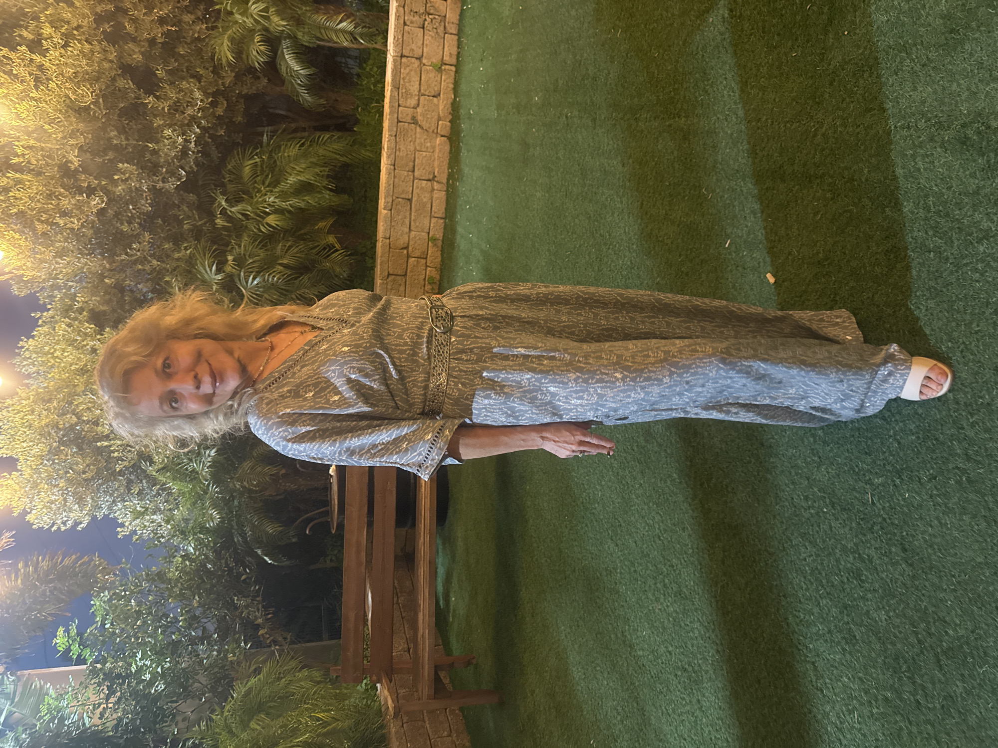 In the city of Migdal HaEmek, Israel, a woman with blonde, curly hair stands outdoors at night. She is wearing a silver, patterned jumpsuit with a decorative belt and white sandals. The woman is standing on a green, artificial turf surface. Behind her, a wooden bench is visible, along with a stone wall and various plants and trees illuminated by overhead lights.