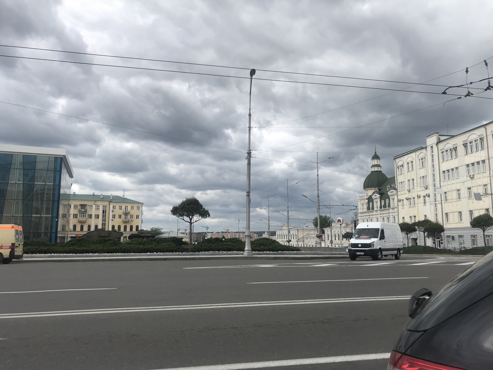 The image shows a street scene in Kharkiv, Ukraine, under a cloudy sky. A multi-lane road stretches across the foreground, marked with white lane dividers. A white van is present on the road, facing the camera. To the left, partially obscured, is an ambulance. In the median, tanks are nestled amongst greenery. Behind the median, there are buildings. A light pole bisects the view. In the background, are historic buildings. The rear end of a vehicle is visible in the lower right corner.