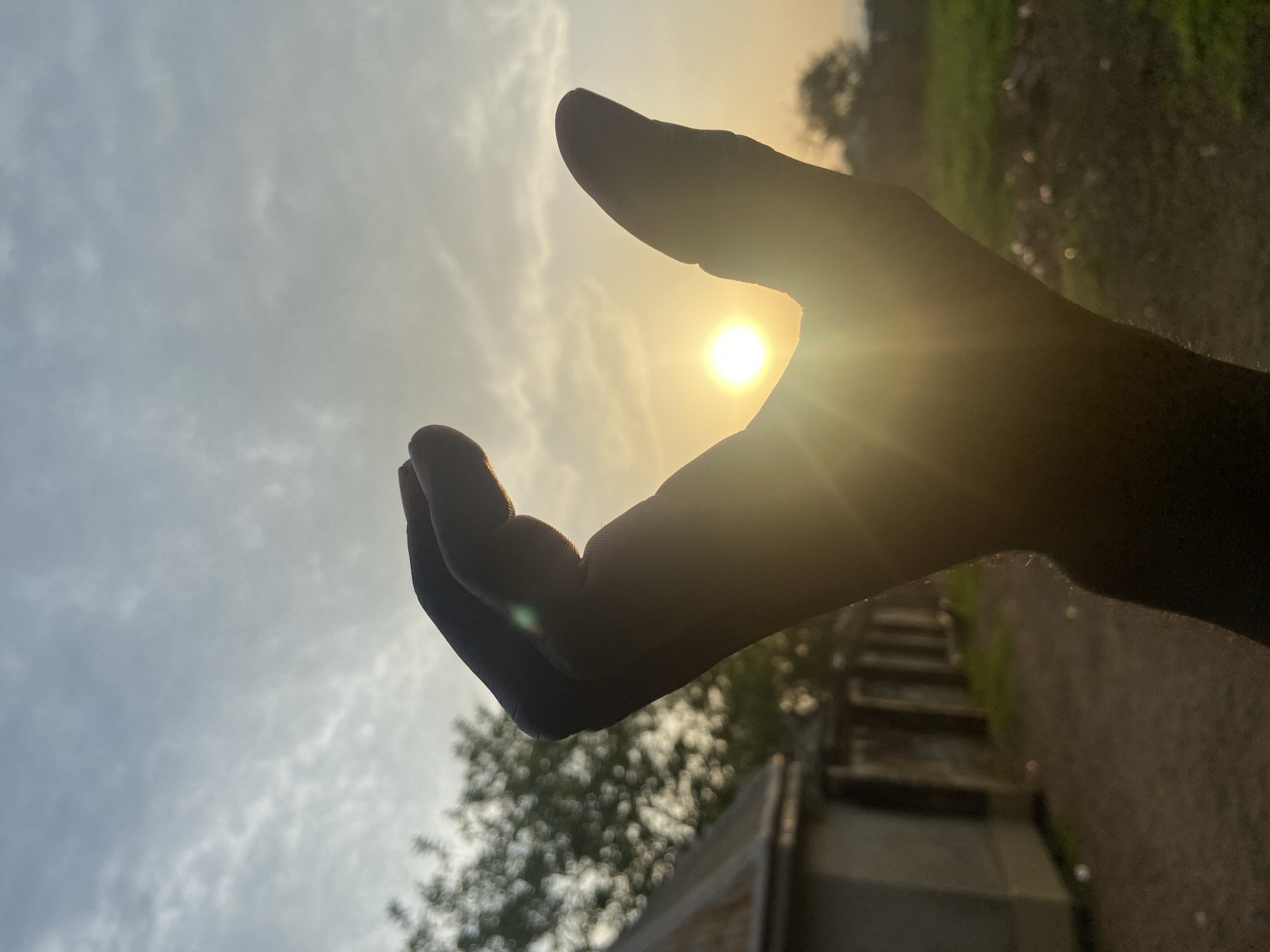 The image shows a human hand silhouetted against the sky, with the sun positioned between the fingers. The background includes a cloudy sky and a blurry view of a building, trees and stairs. The image was taken during daylight hours. No individuals are visible other than the person whose hand is in the frame. There is no explicit evidence to confirm the location is Mararaba, Nigeria.