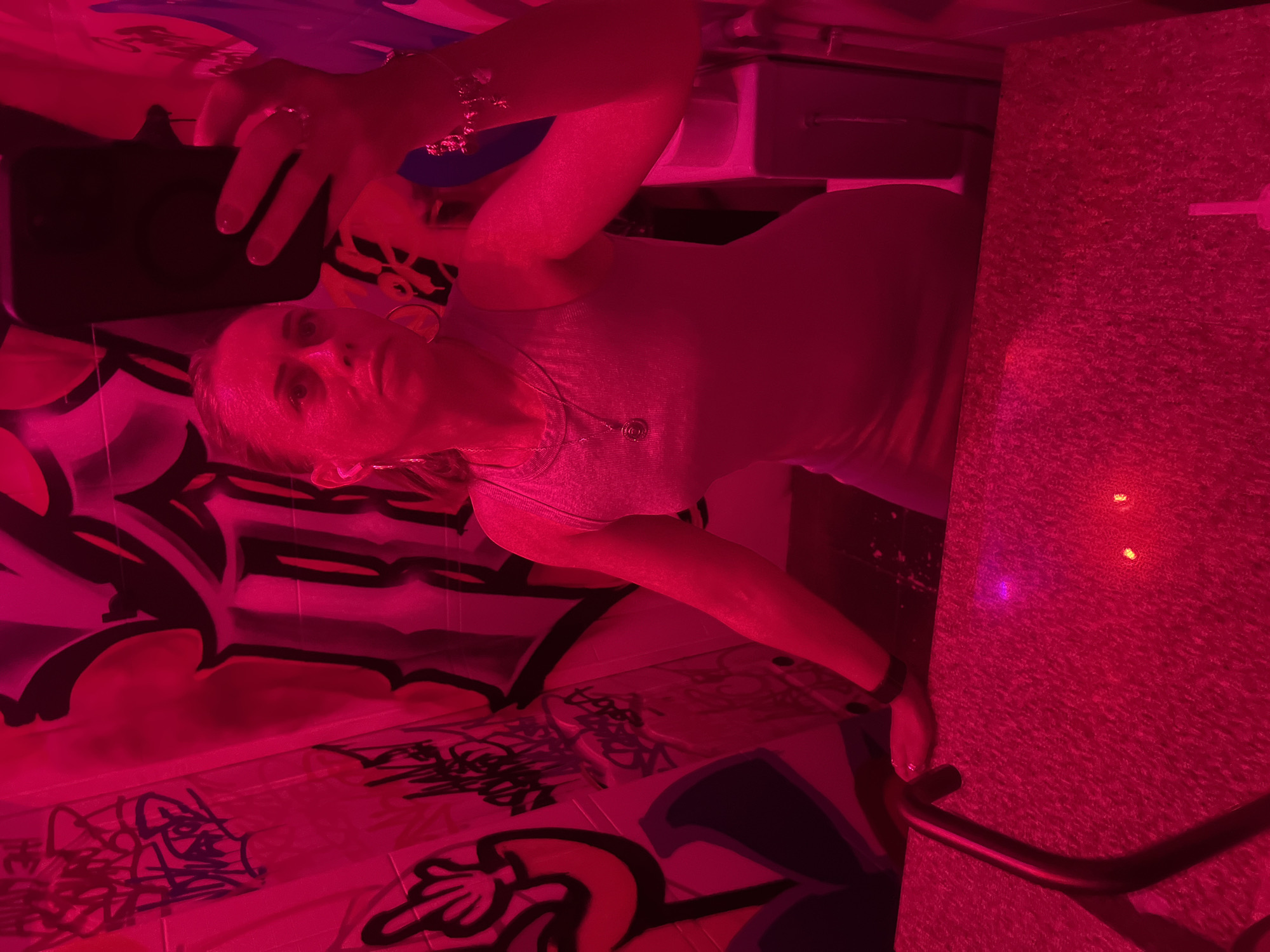 A woman is taking a selfie in a bathroom with graffiti-covered walls. She is wearing a grey sleeveless dress and large hoop earrings. Her hair is pulled back in a ponytail, and she is wearing a bracelet on her right wrist. The lighting is red, casting a pinkish hue over the scene. The wall behind her is covered in colorful graffiti. The floor appears to be made of a speckled material, and there is a black railing in the foreground. The overall impression is that of a vibrant and artistic, albeit somewhat gritty, urban environment. The image location is not specified.