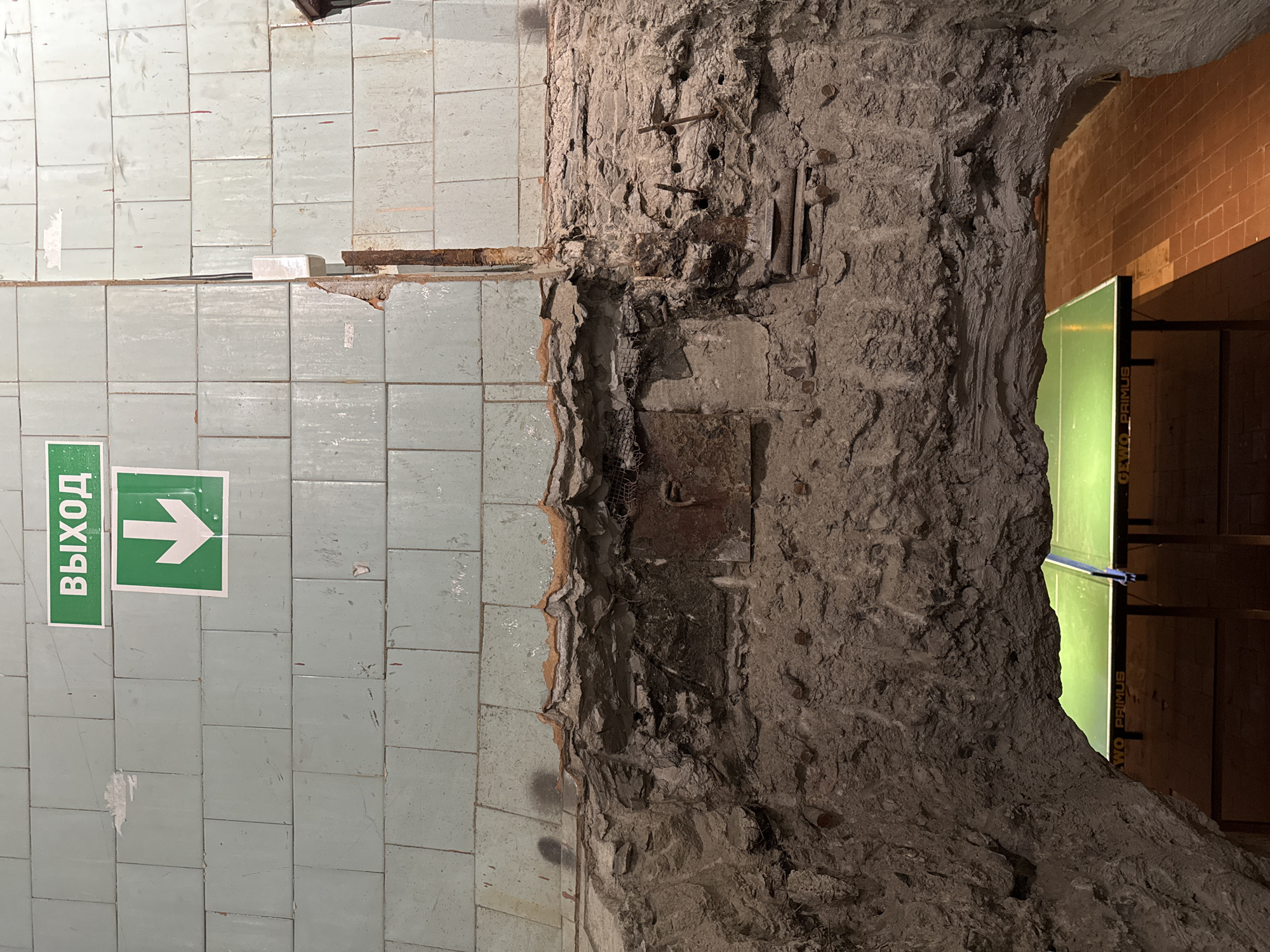 The image depicts a derelict interior space, likely in Saint Petersburg, Russia, based on the presence of a sign reading "выход" (exit) in Russian, situated against a backdrop of pale blue ceramic tiles. The tiled wall is adjacent to a crumbling concrete pillar or wall section exposing rebar and mesh. A corroded metal beam protrudes from the gap between the tiles and concrete. An opening to the right reveals a room with brick walls and a green object, possibly a table tennis table. The overall impression is one of abandonment and decay.