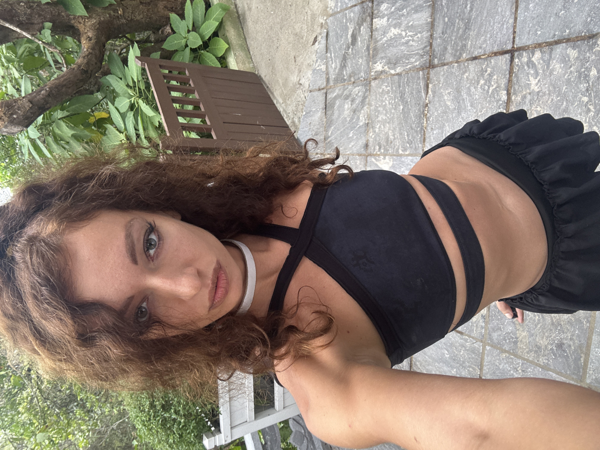 This vertical selfie shows a young woman outdoors in Đà Nẵng, Vietnam. She has long, wavy reddish-brown hair that falls around her face and shoulders, and light green eyes. She is wearing a black strappy top or sports bra that exposes her midriff, paired with black ruffled shorts or a skirt, and a thin white choker necklace. Her expression is direct and composed, looking straight into the camera.
The background features lush green foliage and tree branches, suggesting a natural or garden setting, with a brown wooden slat structure visible on the left side. The ground beneath her is a grey and white mottled stone or tiled pavement. The natural lighting indicates it is daytime. There is no visible text in the image.