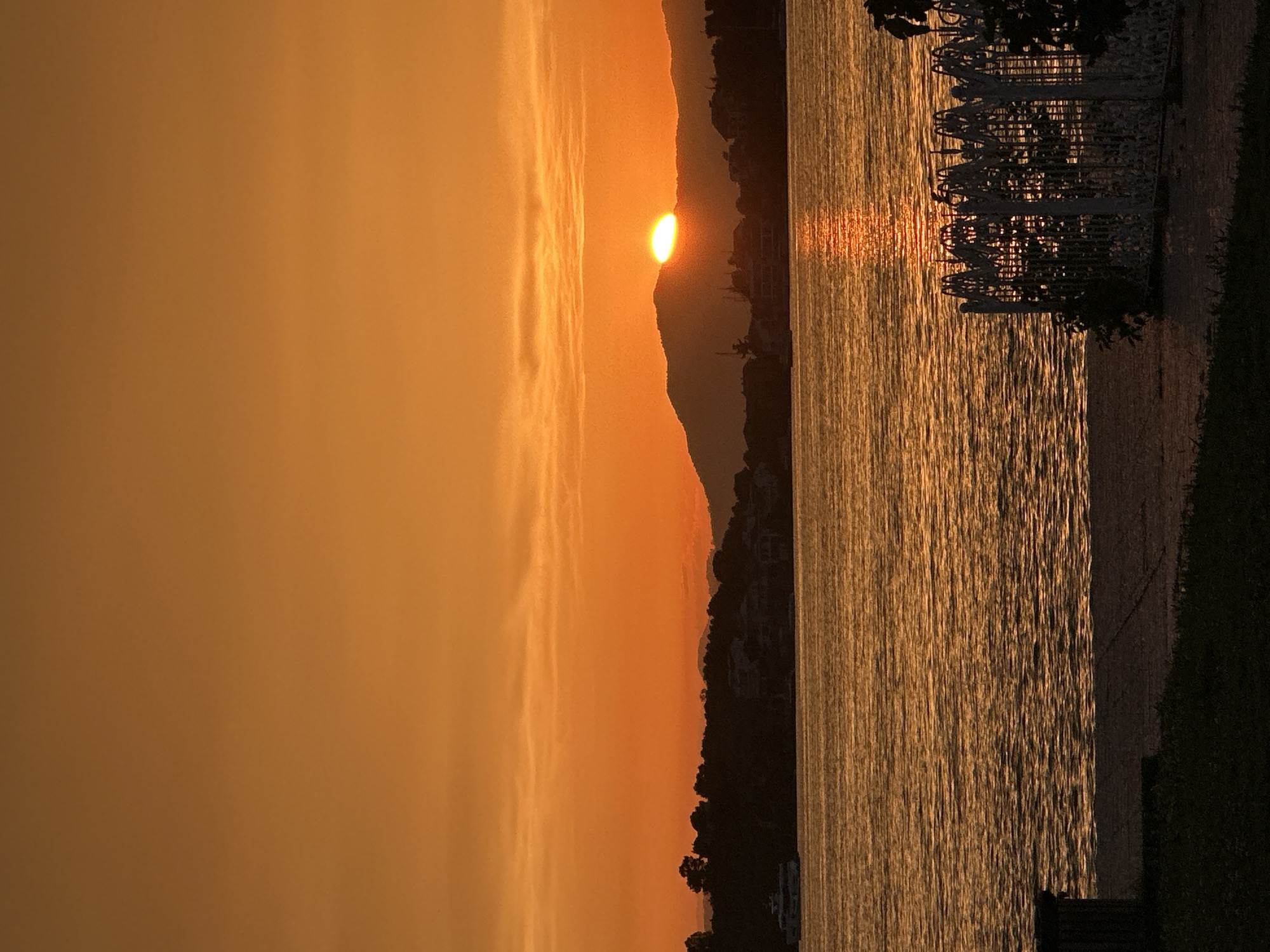 The image depicts a sunset over the water, likely in Fethiye, Turkey. The sun is positioned behind a dark mountain silhouette, its orange glow reflected on the water's surface. A structure with decorative metalwork is visible on the right side of the frame, situated along the shoreline. The sky is a uniform orange hue.