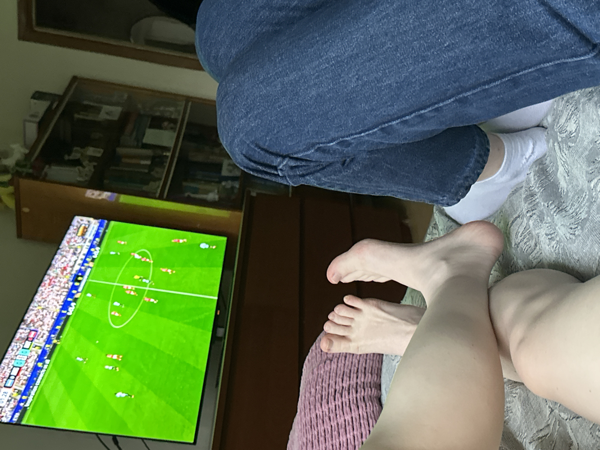 The image shows a first-person perspective of two people lying down, watching a soccer game on a television. The television displays a vibrant green soccer field with cartoonish players. In the foreground, the bare feet of one person are visible, positioned on a patterned surface. The other person is wearing blue jeans and white socks. Behind the television, a wooden bookcase filled with books and decorative items is partially visible, suggesting an indoor setting, possibly a living room or bedroom. The overall ambiance is casual and relaxed, indicative of leisure time. The image does not provide any information about the location in the city of Moscow, Russia.