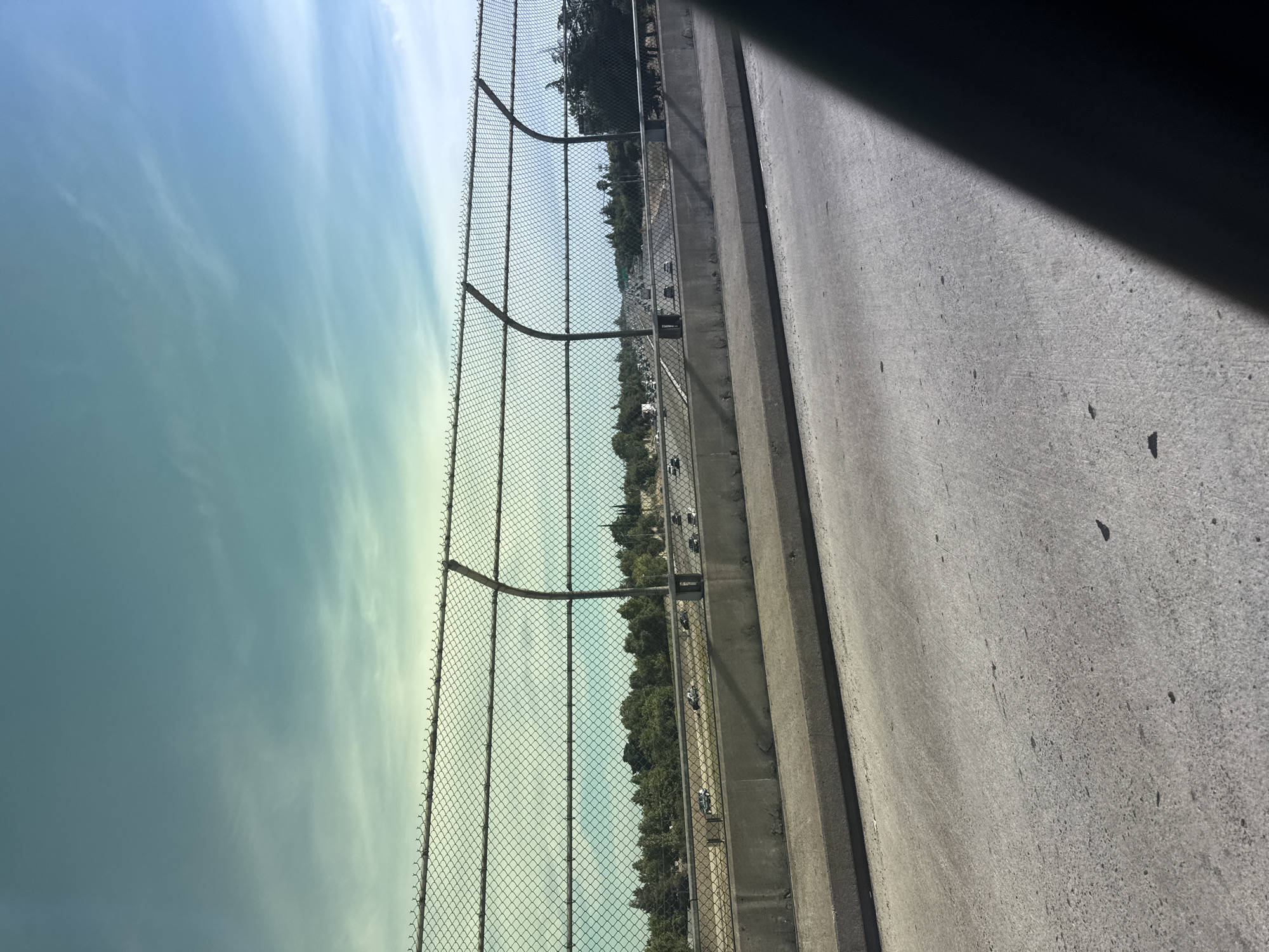 This image captures a view from a raised roadway in Citrus Heights, California, looking out over a multi-lane highway with moderate traffic. A chain-link fence with curved support posts runs along the edge of the roadway, separating it from the highway below. Beyond the highway, a dense line of green trees forms a natural barrier, with some buildings and structures visible in the distance. The sky is a pale blue with wispy white clouds, tinged with a greenish hue due to atmospheric conditions or editing. The foreground shows the concrete surface of the road, with shadows cast by an unseen object. The overall scene depicts a typical urban or suburban freeway environment.