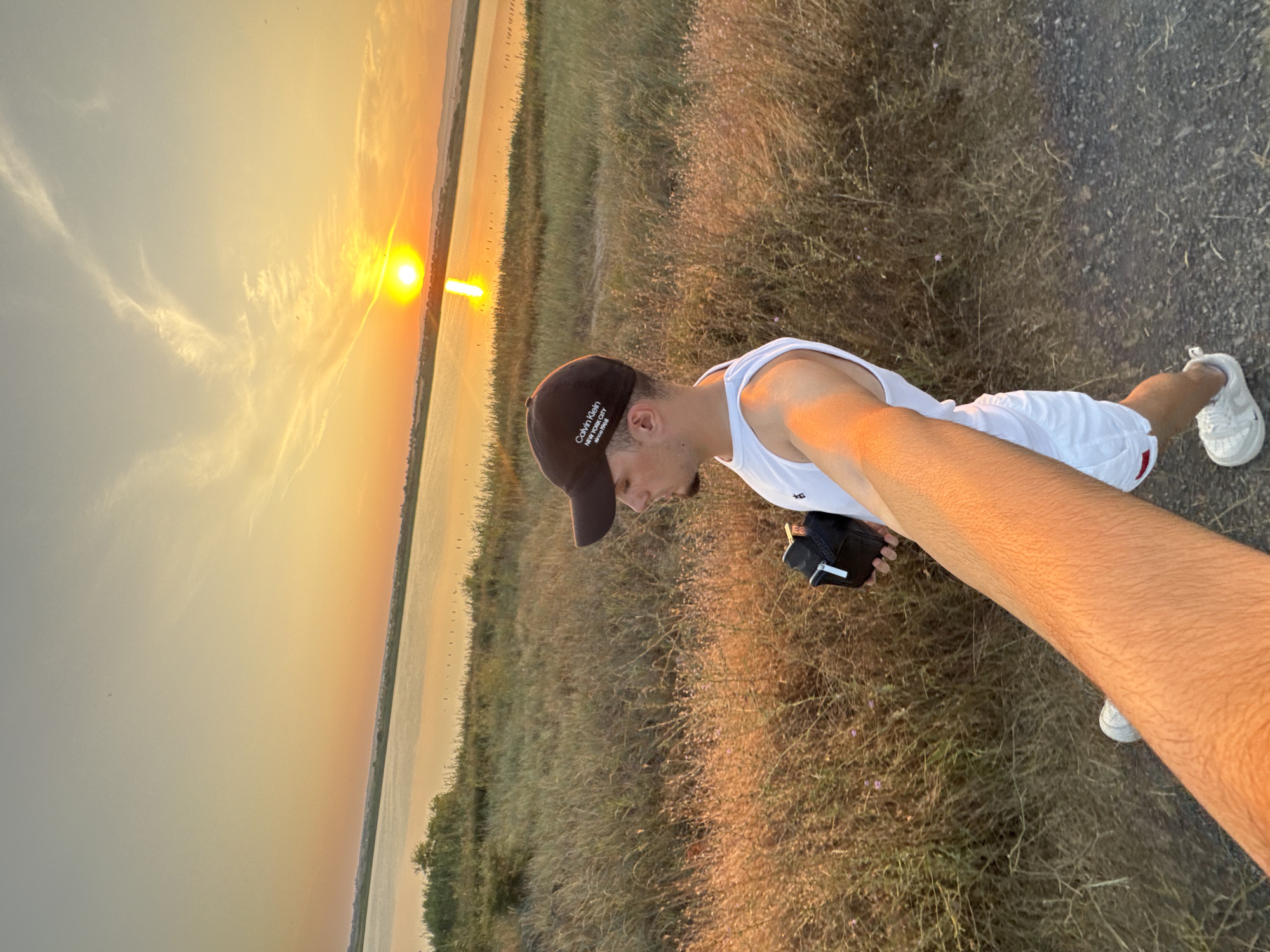 A young man wearing a white tank top, white shorts, a brown baseball cap, and white sneakers is taking a selfie at sunset near the Pomorie salt lake in Pomorie, Bulgaria. He is holding a black camera in his right hand. The sun is setting over the lake, casting a warm orange glow across the sky and water. Tall, dry grass and some bushes are in the foreground. The background shows the salt lake and a distant treeline.