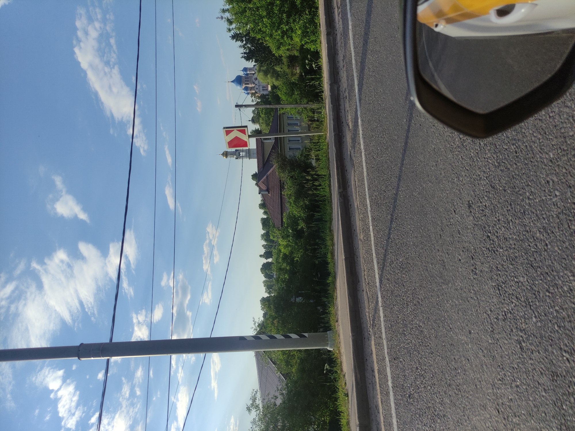 This image depicts a roadside scene in the city of Rogachovo, Russia, under a bright blue sky with scattered white clouds. In the foreground, asphalt pavement of a road is visible, with a white line marking the lane. A sidewalk runs alongside the road, bordered by grass and dense green bushes. A tall, grey utility pole with black and white diagonal markings stands prominently on the left side of the frame.
Further back, a house with a dark, sloped roof is partially obscured by the foliage. A directional sign indicating a turn to the left with a red and white chevron pattern is mounted on a pole. In the distance, a cluster of green trees forms a backdrop. Dominating the far background are two domed buildings, one with a golden dome and bell tower, and another with multiple bright blue domes, characteristic of Russian Orthodox churches. The reflection of a yellow and white object, possibly part of a vehicle, is visible in the lower right corner of the image, suggesting the photo was taken from a car.