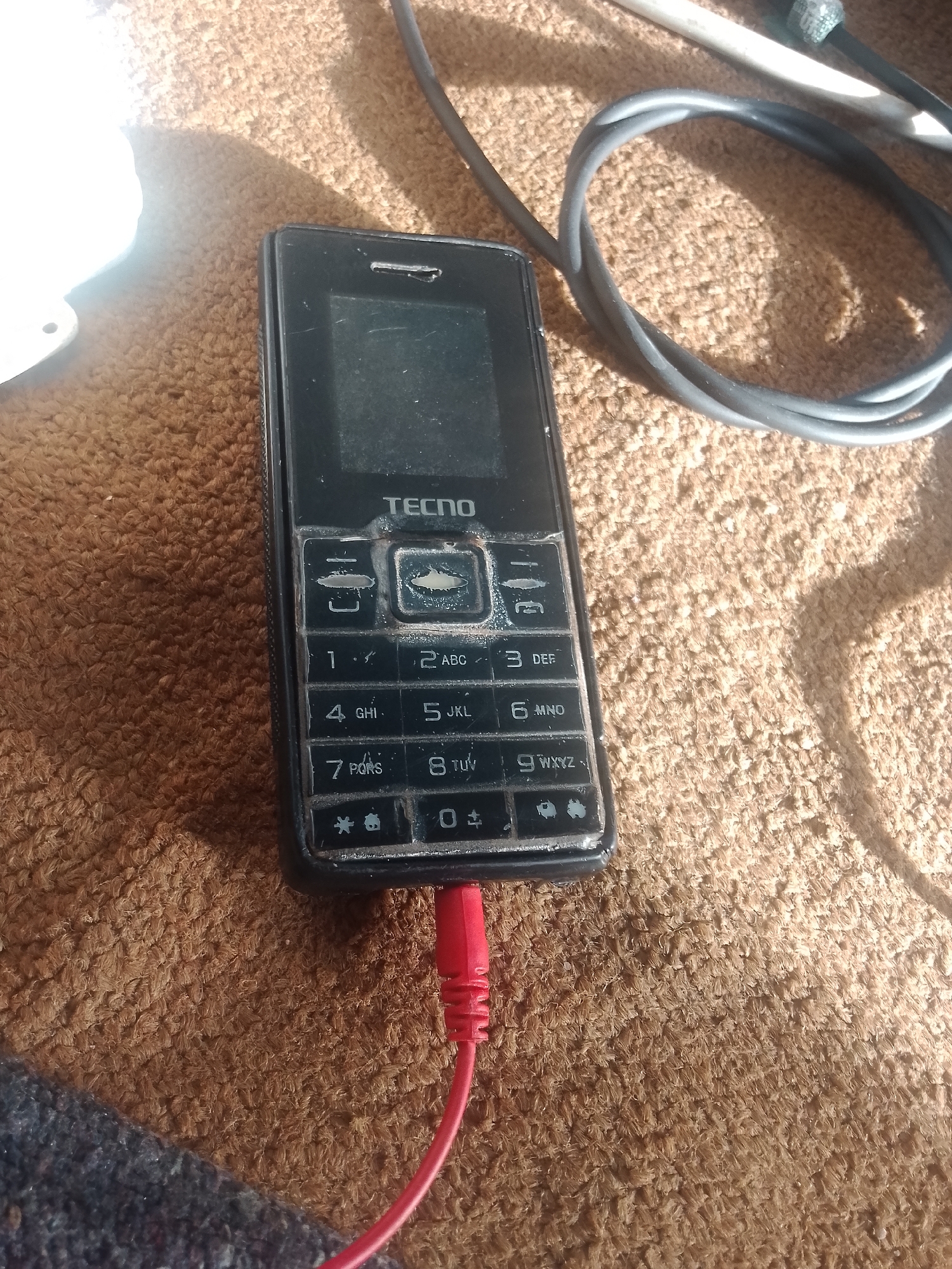 A close-up, high-angle shot shows a black TECNO feature phone lying on a brown, carpeted surface. A red cord, likely a charger or audio cable, is plugged into the bottom of the phone. To the upper right of the phone, a black, coiled cable is visible, and a white cable extends from it. The lighting suggests an outdoor environment, possibly Jalingo, Nigeria, with sunlight casting shadows. The phone's keypad is worn, with some buttons showing significant use. The screen is dark and dusty.