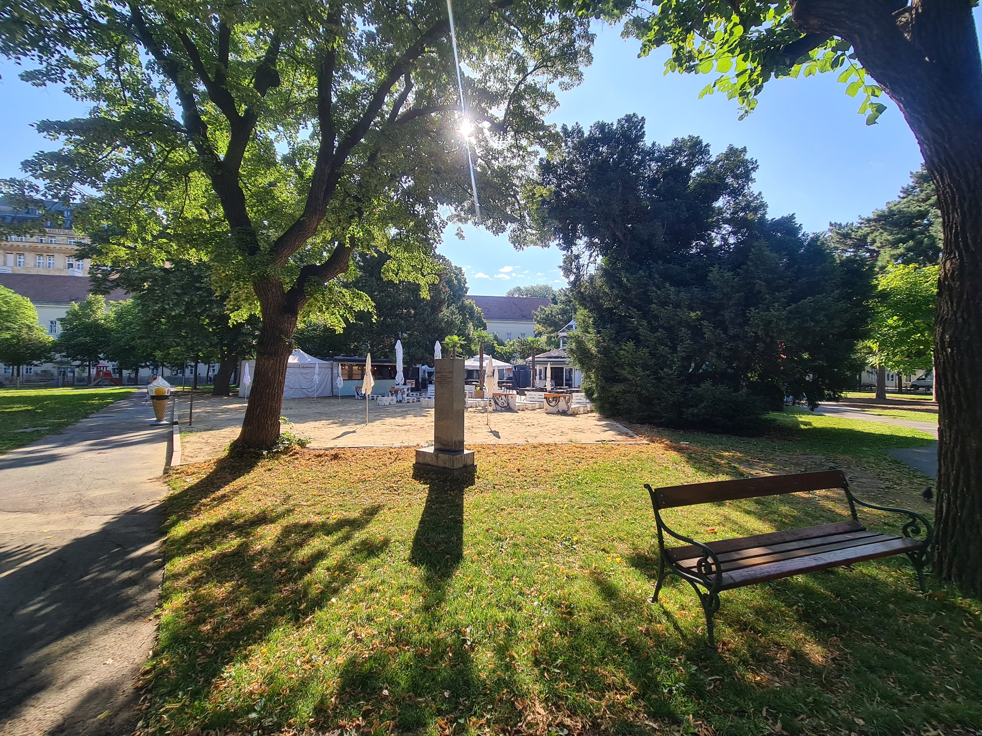 The image depicts a sunny outdoor scene in a park-like setting. In the foreground, a wooden park bench with an ornate metal frame is positioned on a grassy area dotted with fallen leaves. To the left, a paved pathway casts a long shadow from a nearby tree, and a large, stylized ice cream cone sculpture stands beside the path. In the midground, a sandy area contains several white tents, tables, and chairs, suggesting an outdoor seating or event area, possibly a cafe or market. A stone monument stands in the sand. Lush green trees and foliage form a backdrop, with buildings visible in the distance. The sun is shining brightly, casting dappled shadows and creating a lens flare effect. This location is likely a public space within the city of Vienna, Austria, such as a park or square. There are no people visible in the frame.
