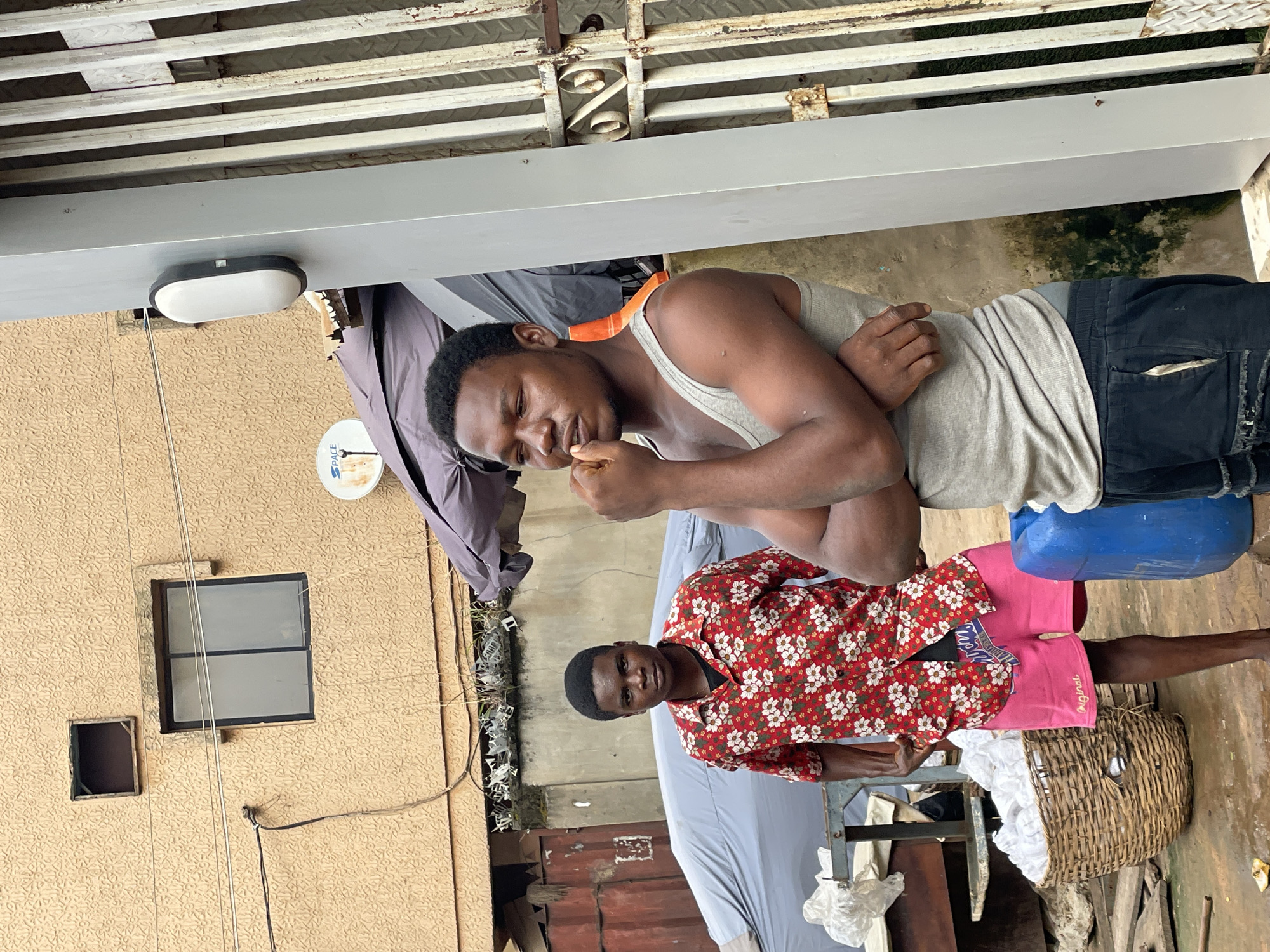 Two men are standing outdoors in an area that appears to be in or near the city of Onitsha, Nigeria.
One man, closer to the camera, wears a grey tank top and dark shorts. He is standing near a blue plastic container. He is holding something in his hand near his mouth.
The other man is wearing a red shirt with a white floral pattern and pink shorts. He is standing beside a wicker basket filled with white fabrics. The scene includes a building, a satellite dish, laundry hanging, and other outdoor objects indicating a residential or commercial area. A metal container is also present in the background.