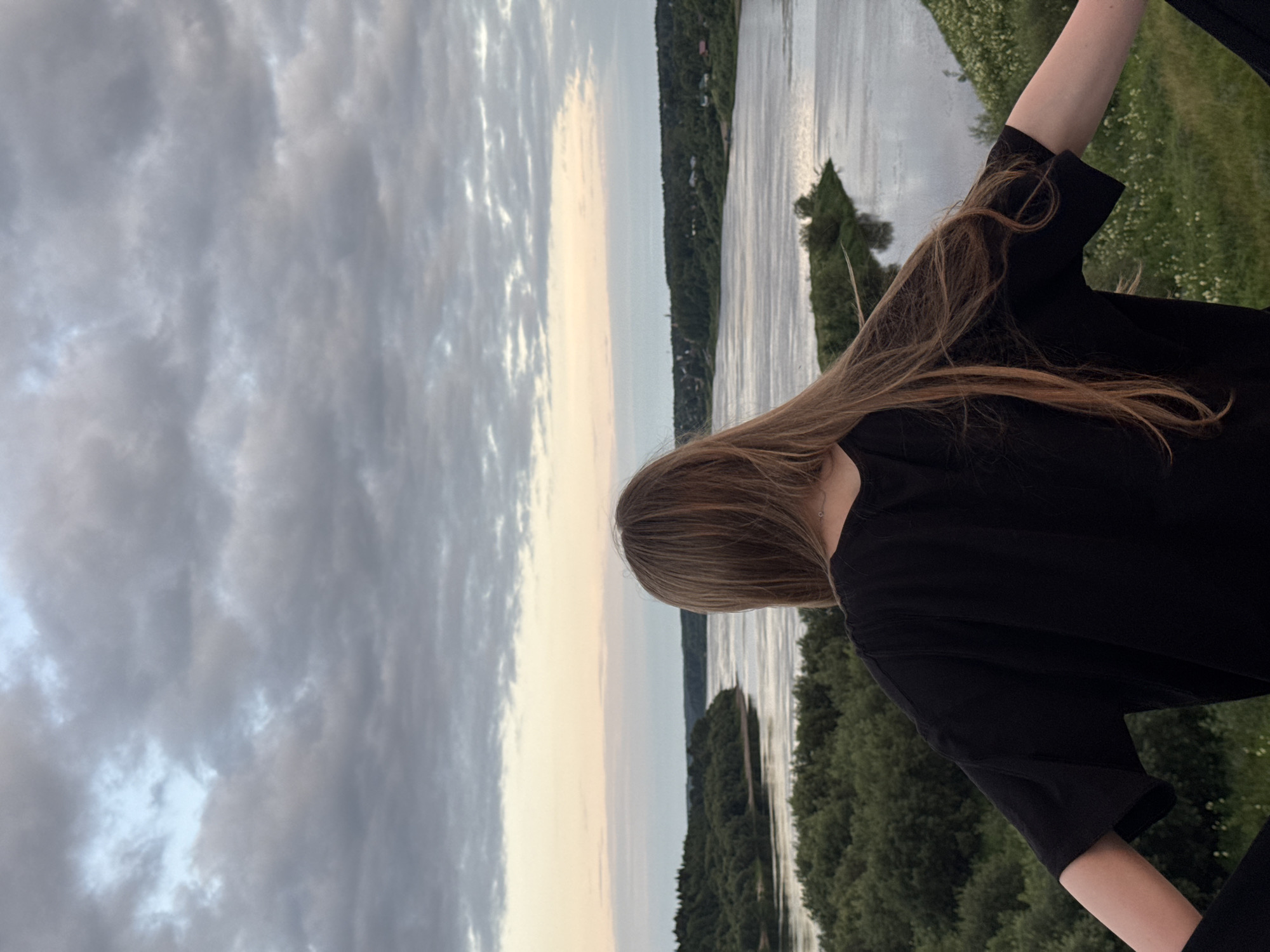 A person with long, blonde hair is viewed from behind, facing a wide river and a lush, green landscape under a cloudy sky. The person is wearing a black t-shirt and is positioned on a grassy embankment overlooking the river. The river curves through a densely wooded area, with hints of buildings visible on the distant shore. The sky is filled with soft, diffused light from a sunset or sunrise, with pastel hues of pink and orange visible near the horizon. The overall scene suggests a tranquil natural setting, possibly in the village of Ryabinino, Russia, known for its scenic river views.