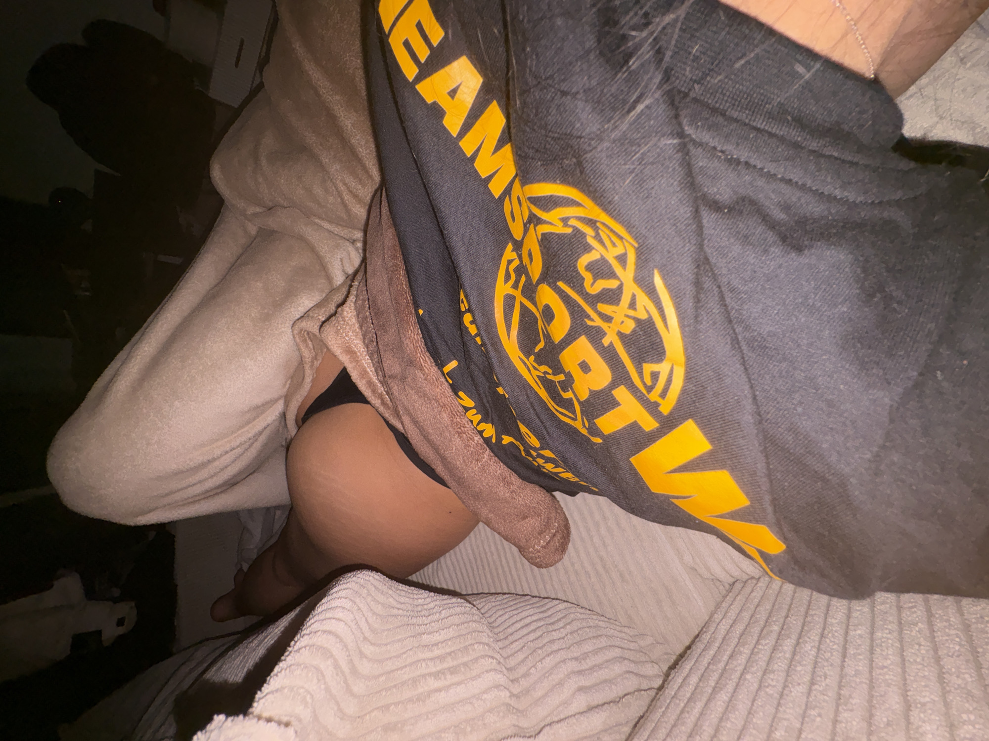 The image is a close-up, low-angle shot of a person lying down. The person is wearing black underwear and a dark t-shirt with a yellow graphic design. A tan towel is draped over their body. The setting appears to be indoors, possibly a bedroom, with a textured light-colored surface to the left of the frame, likely a sofa or bed. The lighting is dim, creating shadows. There is no indication of the city of Mönchengladbach, Germany, or any other specific location.