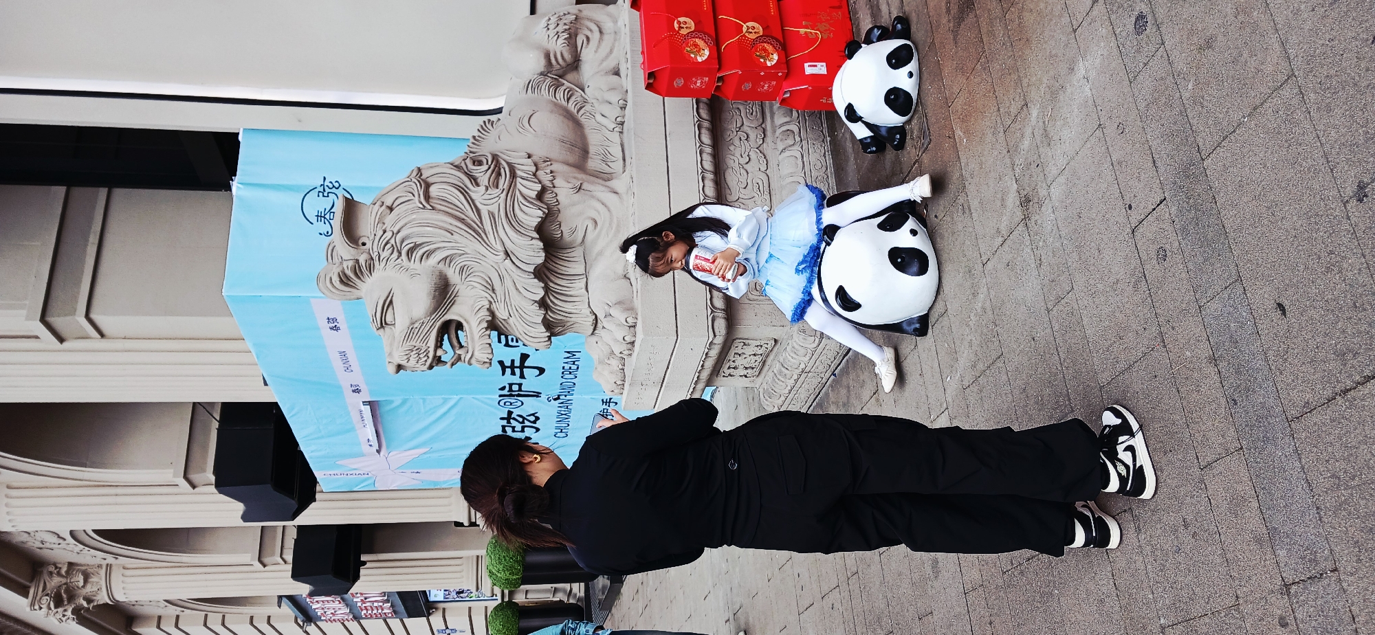 A medium, eye-level, rotated outdoor shot shows a woman taking a photograph of a girl posing with two panda statues in the Yuzhong District, China. The woman wears a black top and pants, and white and black sneakers. She holds a cell phone up to take the picture. The girl poses in a light blue dress and white leggings. She sits on top of the larger panda statue and holds a small red container in her hands. Behind the panda statues are red packages with tassels. To the left of the subjects is a display that has the sculpted head of a lion. The banner on the display reads, "CHUNXIAN HAND CREAM."