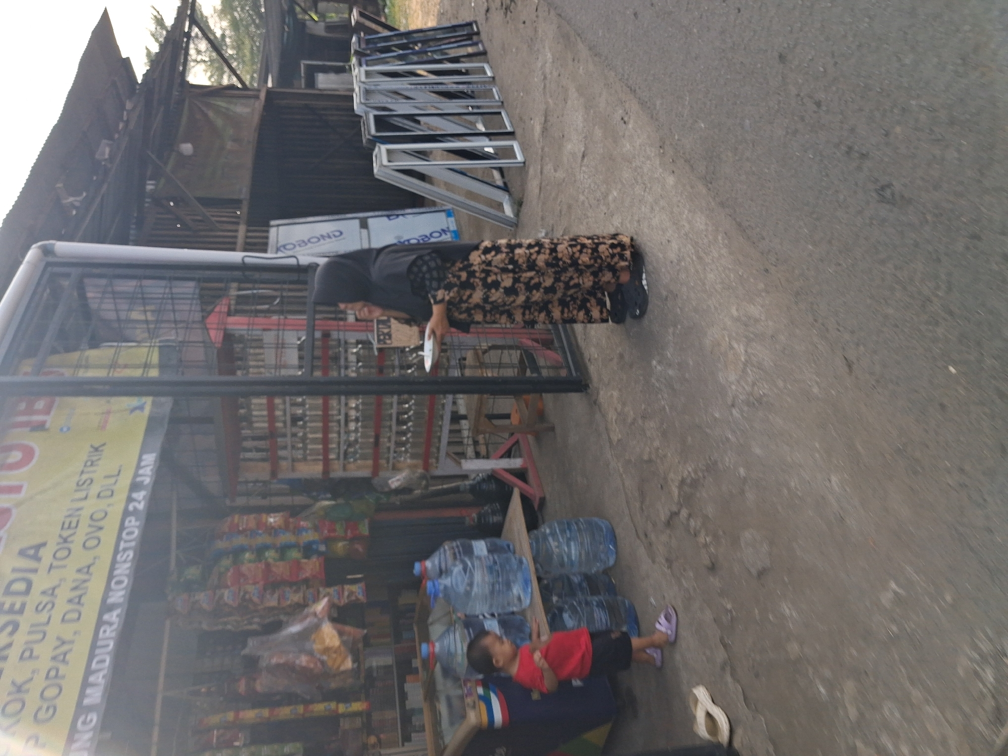 In Bojong Gede, Indonesia, a young child in a red shirt and black shorts stands near several large blue water jugs. A woman wearing a black hijab and a patterned long skirt is standing to the right of the child, holding a white plate. She is looking towards the child. In the background, a storefront displays a banner with Indonesian text. The area appears to be an outdoor market or roadside stall, with various items for sale including packaged goods hanging from racks and what appear to be window frames stacked on the ground. The ground is paved with concrete and asphalt. The lighting suggests it is daytime.