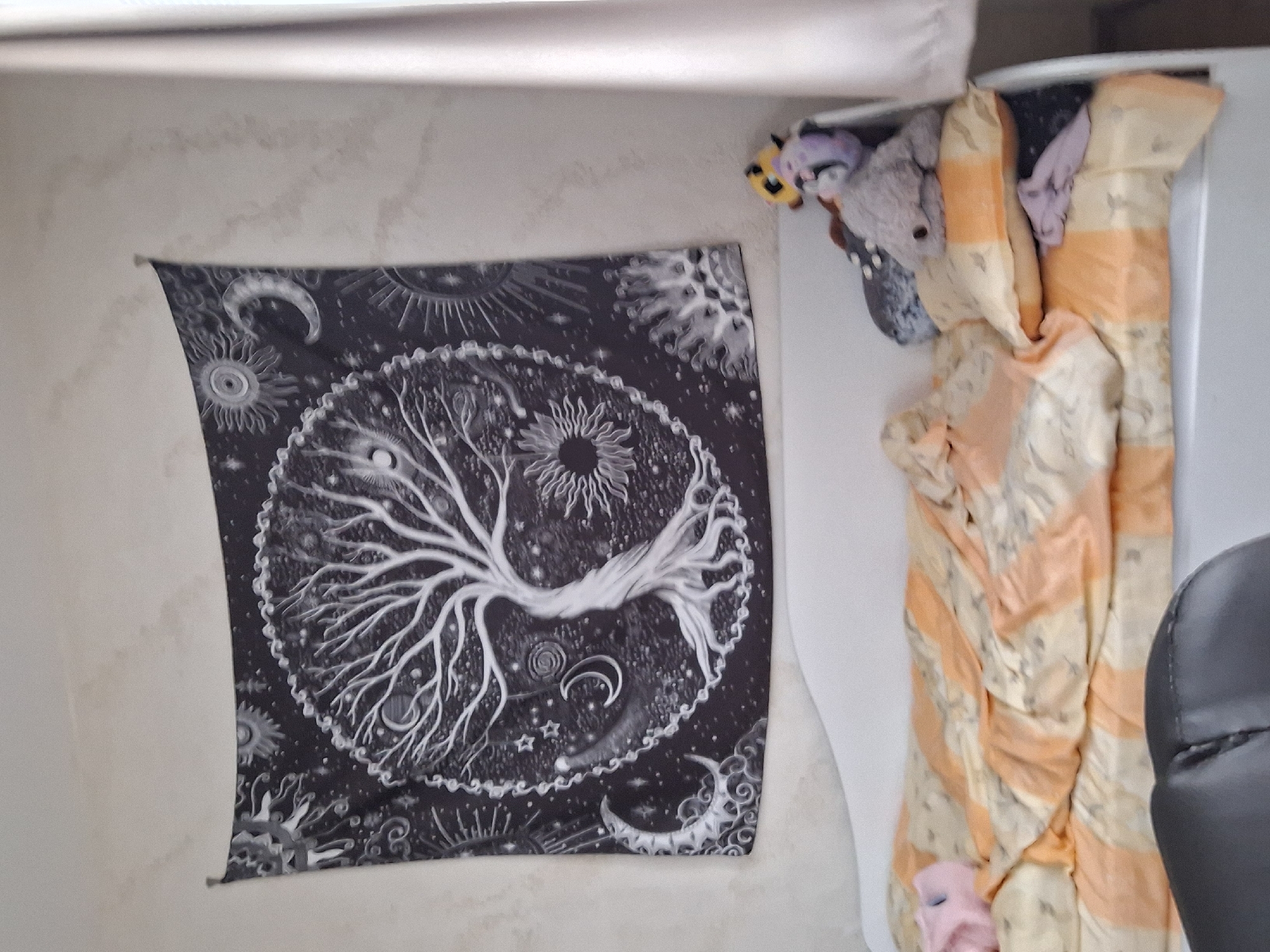 The image presents an indoor scene. On the left, a wall is adorned with a black and white tapestry featuring a prominent tree design, surrounded by celestial motifs. To the right, a partially obscured area reveals a cream-colored textile with orange stripes, interspersed with soft toys including a grey plush bear and what appears to be a pink plush creature. The right edge of the frame shows a portion of a dark-colored office chair. The environment suggests a room in a domestic setting. There is no evidence to suggest the location is in Panagyurishte, Bulgaria.