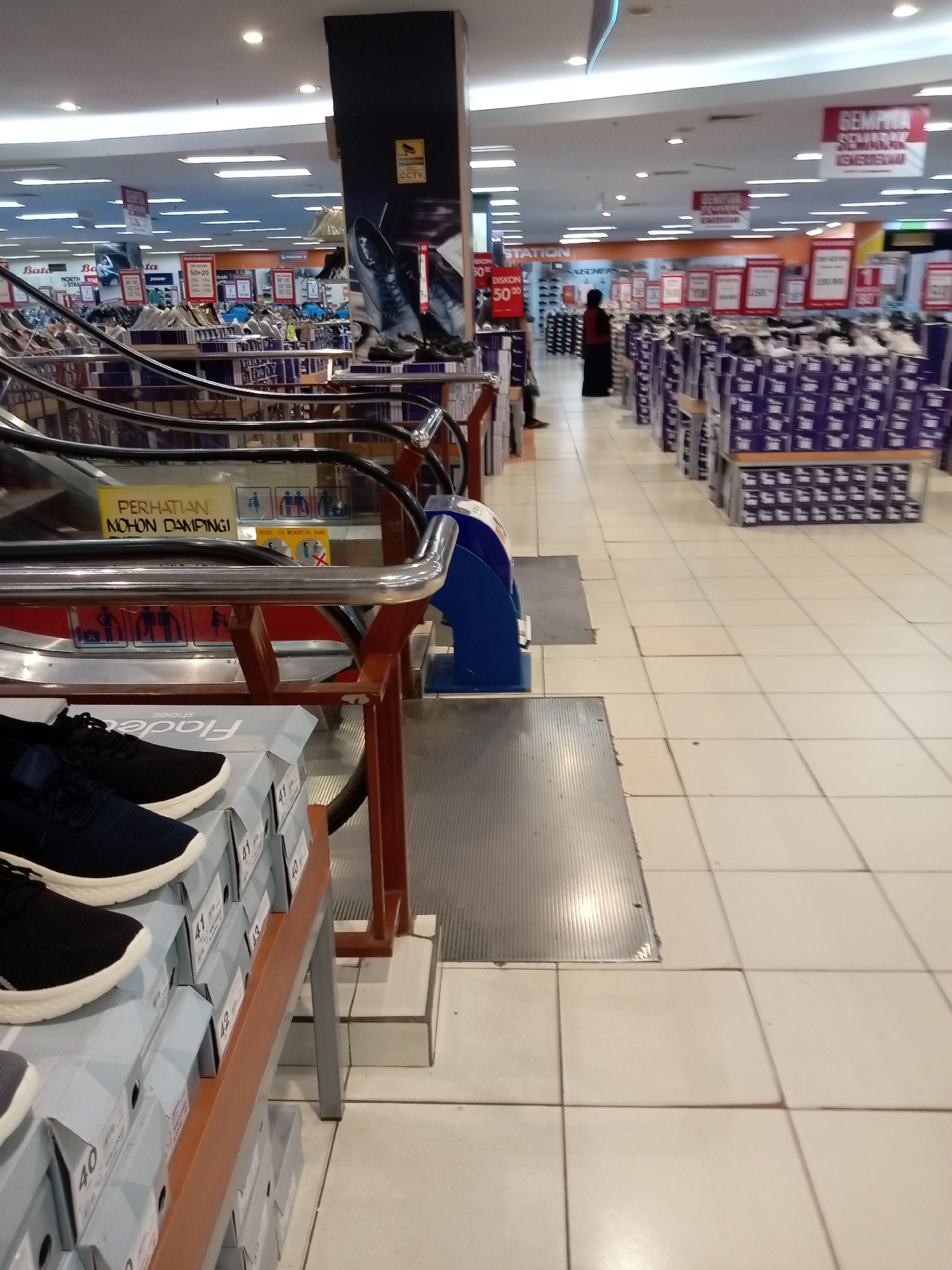 The image captures an indoor scene within a department store, likely in Batam, Indonesia, showcasing a shoe retail section. In the foreground, a stack of shoe boxes is visible, with two pairs of athletic shoes, one black and one navy, placed on top of them. Adjacent to this display is a section of an escalator with dark metal railings. Further into the store, numerous shelves are filled with more shoe boxes, predominantly colored purple and white. Signs indicating discounts, such as "DISKON 50%" (50% discount) and "DISKON 30%" (30% discount), are prominently displayed. One sign also reads "GEMPITA SEMARAK IGNEUSIUSI," which translates to "Ignite the Festive Spirit." A woman in a maroon outfit and dark skirt is seen walking away from the camera towards the back of the store. The floor is tiled with light-colored square tiles. Above, fluorescent lighting illuminates the vast retail space, which appears to be a large shopping mall or a hypermarket.