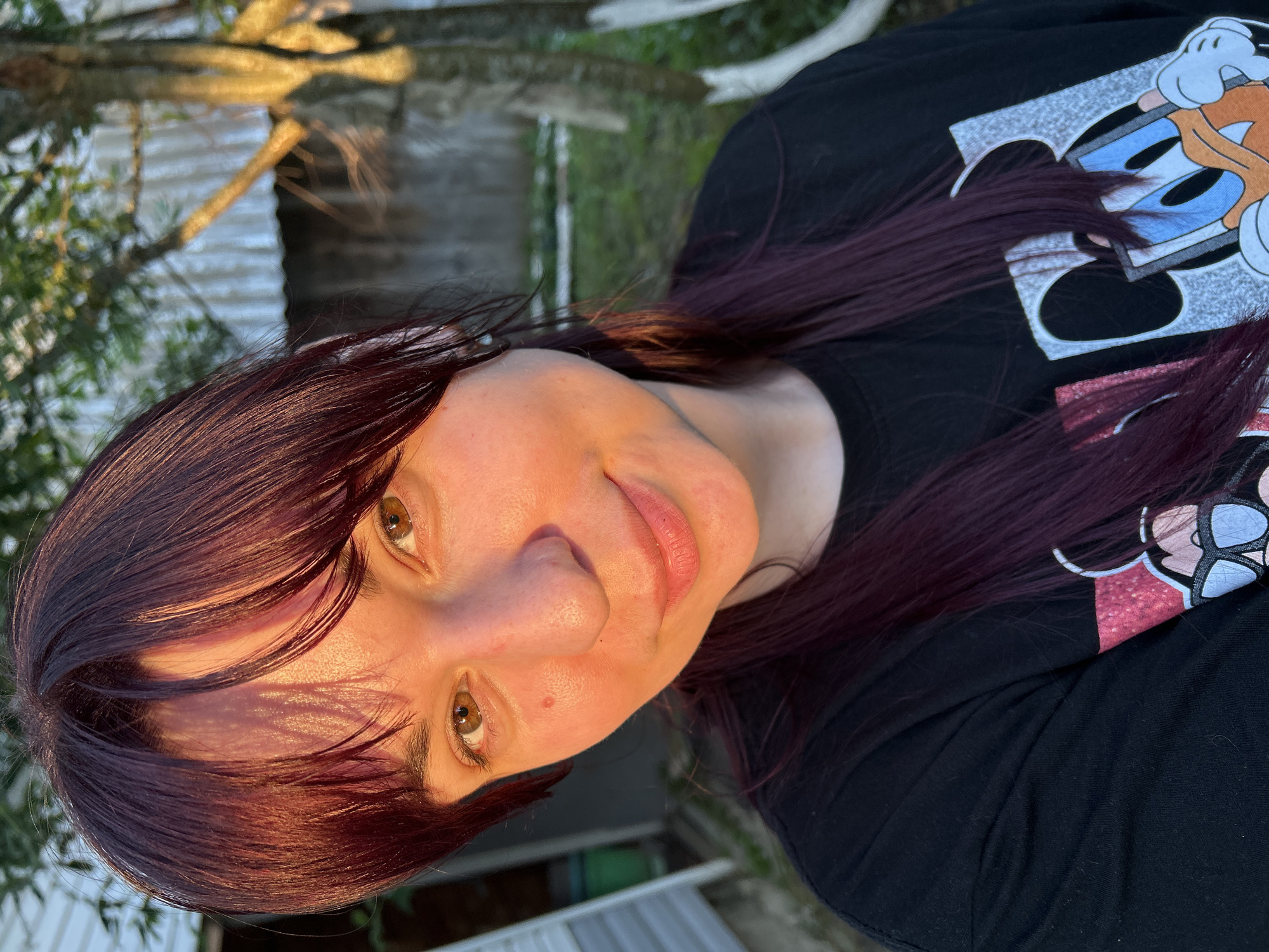 The image shows a close-up, outdoor, eye-level shot of a young woman with long, dark, burgundy hair. She is wearing a black t-shirt with a graphic print of Donald Duck. The woman is looking towards the camera with a slight smile. The lighting suggests it is either early morning or late afternoon, with warm sunlight illuminating her face and hair. The background is blurred but appears to be a backyard setting with greenery and a fence. There is no indication of the specific location in the city of Фоки, Russia.