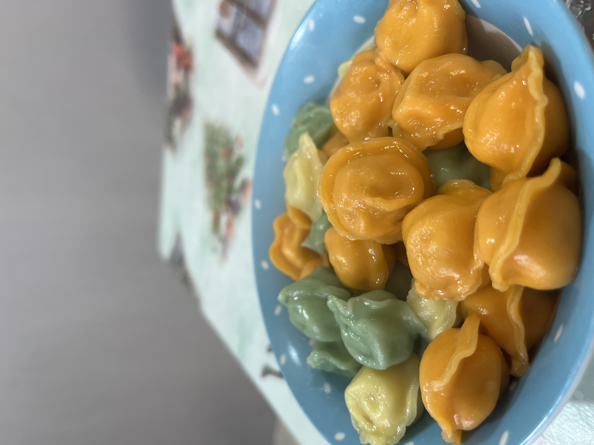 The image displays a close-up, slightly tilted view of a light blue bowl filled with a variety of colorful dumplings. The bowl has small white polka dots and is placed on a light-colored, patterned tablecloth. The dumplings are glossy and appear freshly cooked, with dominant hues of bright orange and yellow, interspersed with some light green and pale yellow pieces. They are small, plump, and intricately folded, resembling a type of pasta like tortellini or pelmeni.
To the left of the bowl, a blurred, plain grey surface, possibly a wall or another table, occupies a significant portion of the frame. The patterned tablecloth, visible beneath and around the bowl, features faint, stylized designs that include what appears to be a cactus-like plant and architectural elements.
The scene is an indoor setting, likely a kitchen or dining area, focused on a prepared meal. There is no discernible activity taking place. Given the location context of Chaykovsky, Russia, these dumplings are consistent with popular local dishes such as pelmeni or vareniki. No specific time of day, weather conditions, or emotions are conveyed in the image, and there is no visible text.