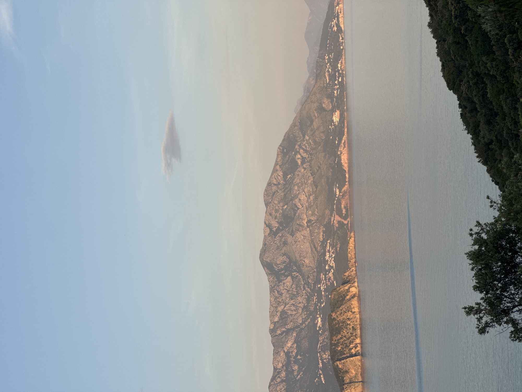 The image depicts a landscape with a mountain range running along the center. The left portion of the image is filled with a light blue sky containing a single wispy cloud. To the right of the mountain range is a large body of water, with dense green vegetation visible along its rightmost edge. The mountain range appears to be illuminated by sunlight, accentuating its rugged texture. There are no people or discernible actions visible. Based on the location of Dubraljevina, Montenegro, the body of water is likely the Adriatic Sea.