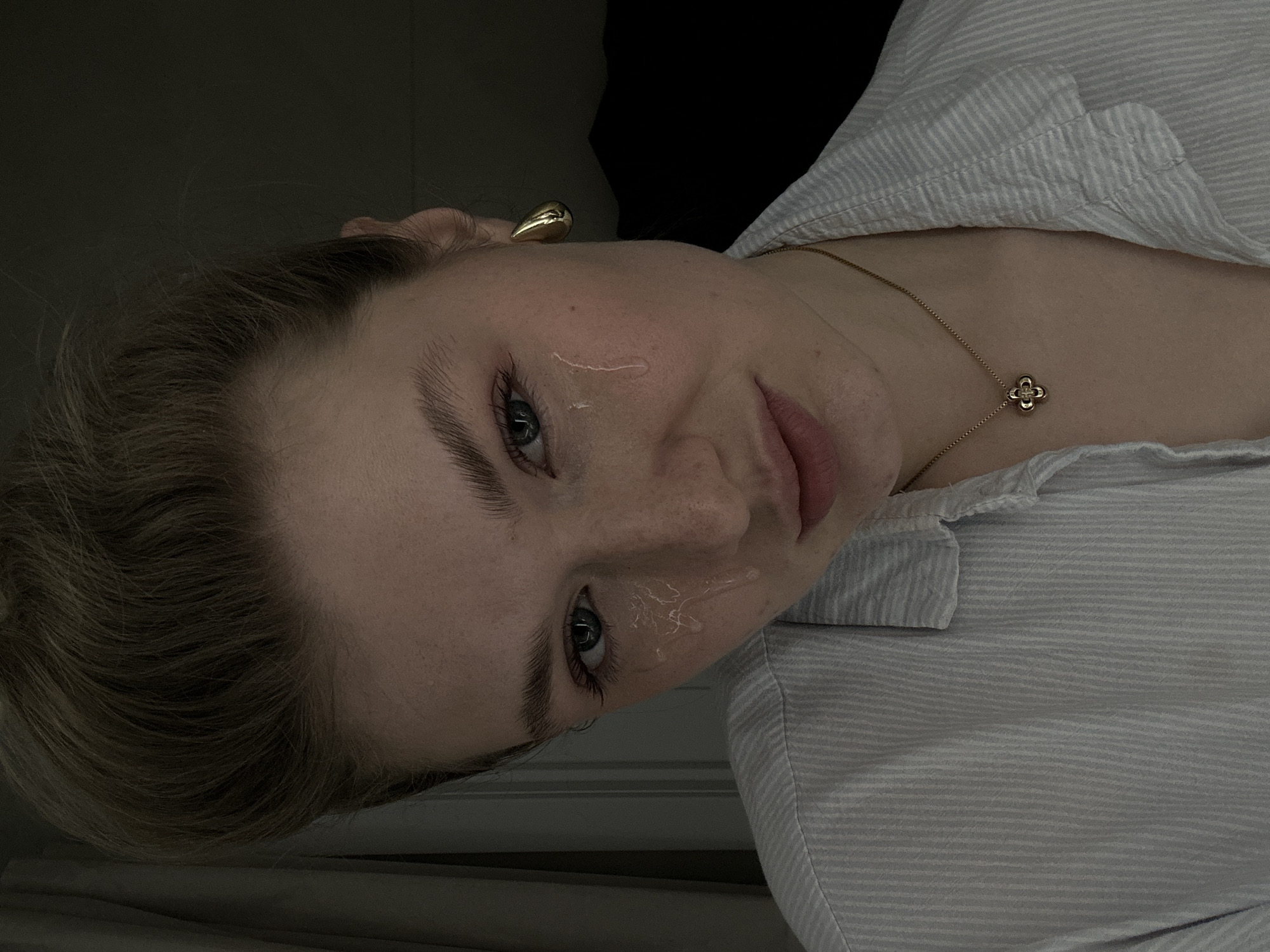 A close-up, slightly canted, indoor photograph shows a fair-skinned woman with light blonde hair and blue-grey eyes. She is wearing a light-blue and white striped shirt. A thin gold chain with a clover-shaped pendant is visible around her neck. She has a gold earring in her left ear. Two clear, liquid streaks are visible on her face. The background is a dark, undefined space. There is no information suggesting the location is Kazan, Russia.