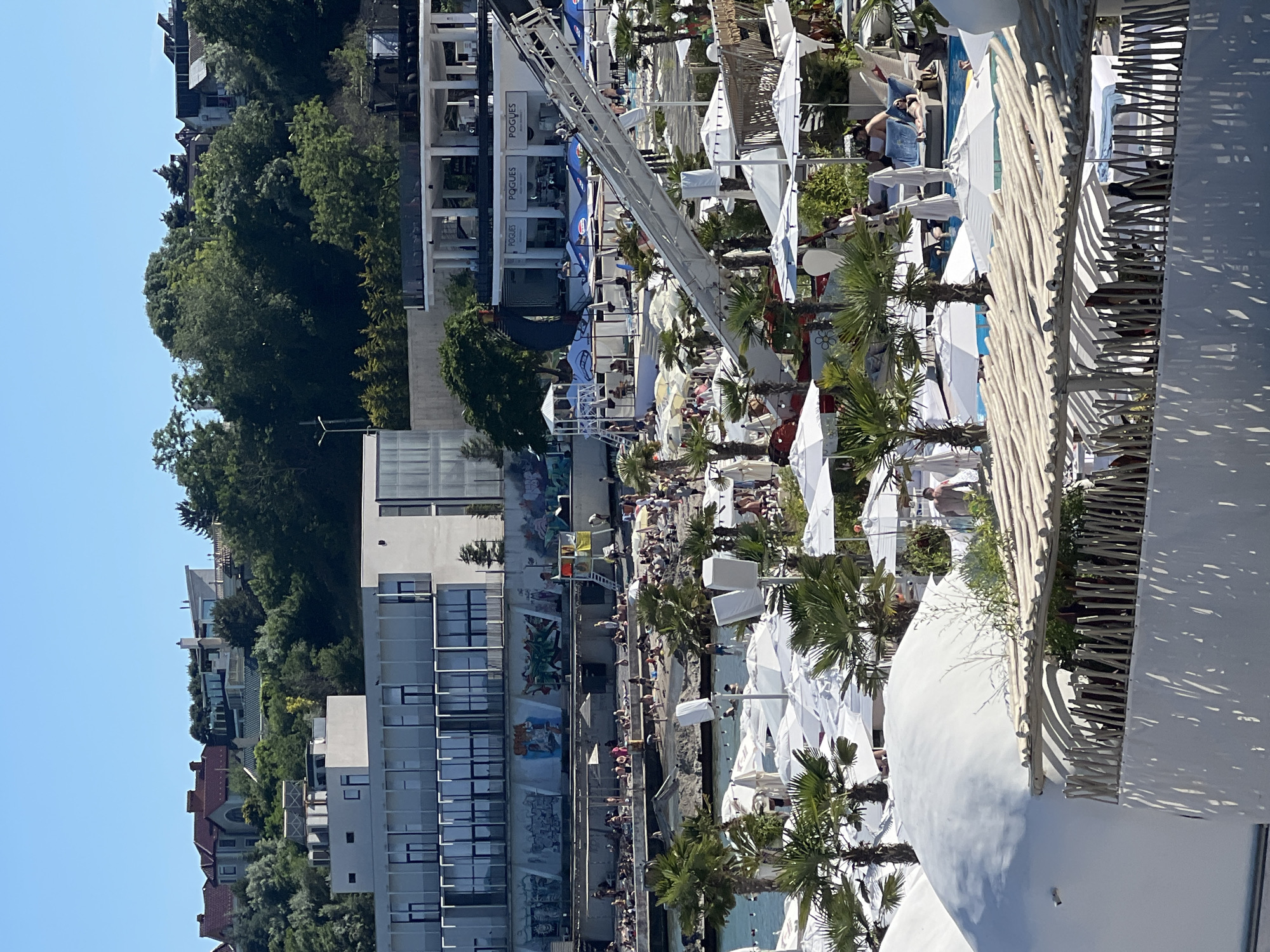This is a wide shot of a crowded beach resort in Odesa, Ukraine, on a clear, sunny day. In the foreground, a white, domed structure and a rustic, wooden-roofed cabana are visible. Beyond them, a bustling beach scene unfolds, with numerous white umbrellas, lounge chairs, and people relaxing and swimming in the clear blue water. Palm trees dot the landscape, adding to the tropical feel. In the background, a modern white building with large glass windows stands against a backdrop of lush green trees and residential buildings on a hill. The scene captures the vibrant atmosphere of a popular seaside destination.
