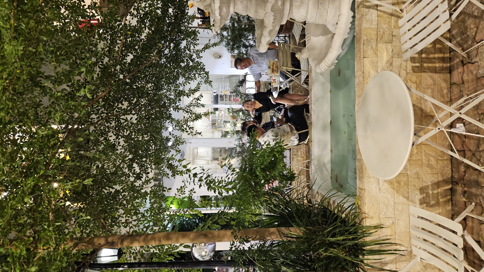 The image shows an outdoor cafe scene in Nha Trang, Vietnam. Several people are seated at tables, engaged in conversation. A white, round table and matching chairs sit on a paved surface, possibly brick or stone. Nearby is a narrow water feature or shallow pond, lined with similar stone or brickwork. Lush greenery, including trees and plants, surround the seating area. A lamppost with a globe-shaped fixture is visible among the foliage. The background suggests the cafe extends indoors, visible through large glass doors or windows. A building is observed in the background.