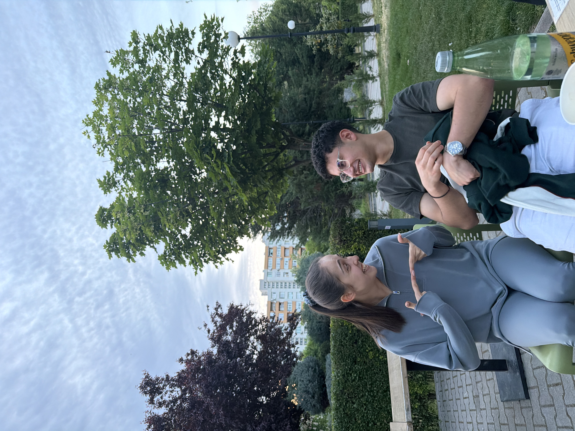 Two people are seated at a table outdoors. A man with dark hair and glasses is wearing a dark t-shirt and a watch and holding a green jacket. He is smiling at a woman, dressed in a gray tracksuit, who is smiling back at him and holding up her hands with her index fingers and thumbs extended, forming a "L" shape with each hand. A bottle of Schweppes and a white bowl are on the table. Behind them are trees, a lamp post, a hedge, a building, and a cloudy sky. The scene appears to be in a park in Blagoevgrad, Bulgaria.