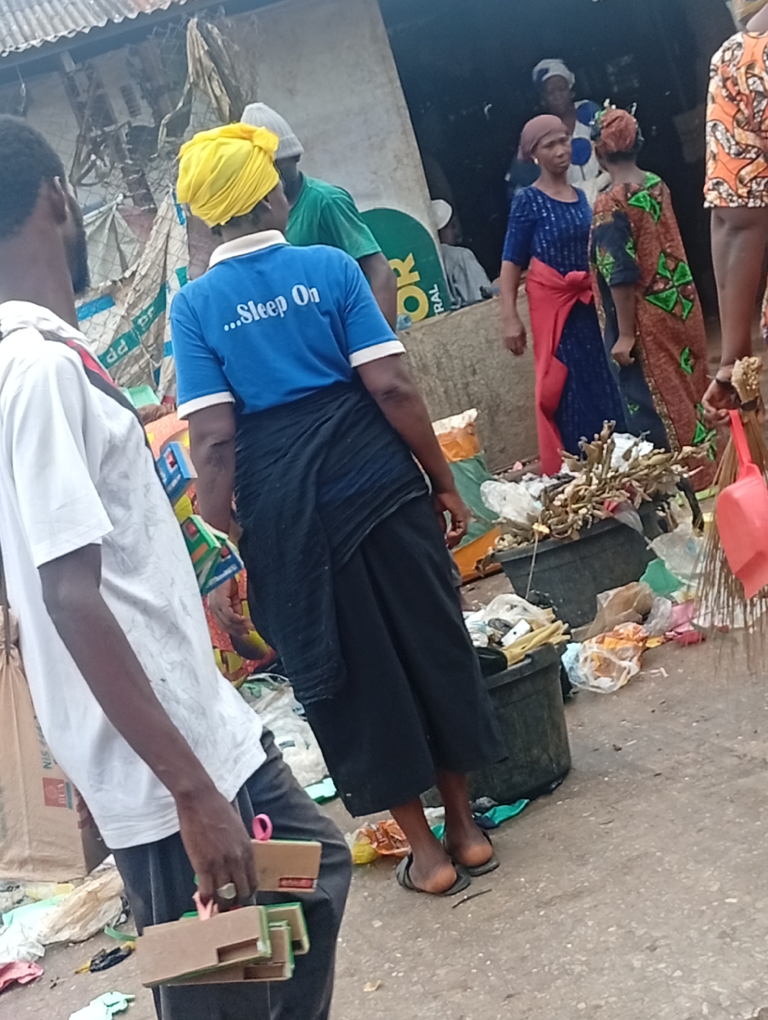 The image shows a street scene in Maiburiji, Nigeria. There are several people present, including a man in a white shirt holding cardboard objects, a woman with a yellow headwrap wearing a blue shirt that says "...Sleep On," and several other individuals in traditional clothing. The environment is littered with trash. A woman is holding a broom and dustpan. The scene is a busy market or street, with open-air businesses visible in the background.