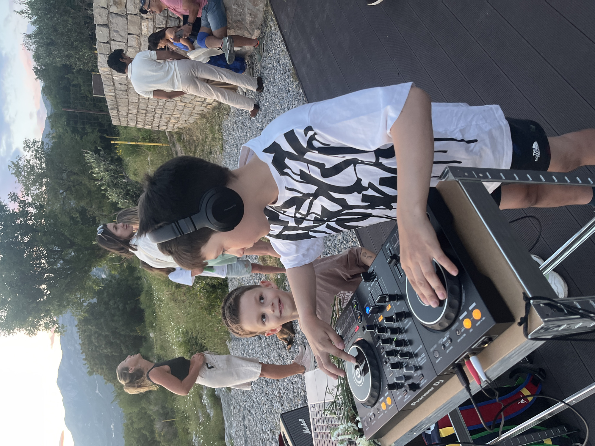 A DJ setup is on an outdoor deck, likely in a backyard, with a Pioneer DJ controller on a stand, and a Marshall speaker visible. A young boy wearing headphones is operating the controller, while another child is looking at the camera. Several people are standing and sitting in the background, in an area with gravel and grass, with a mountain landscape in the distance. The location is likely Vojnići, Montenegro, based on landscape features.