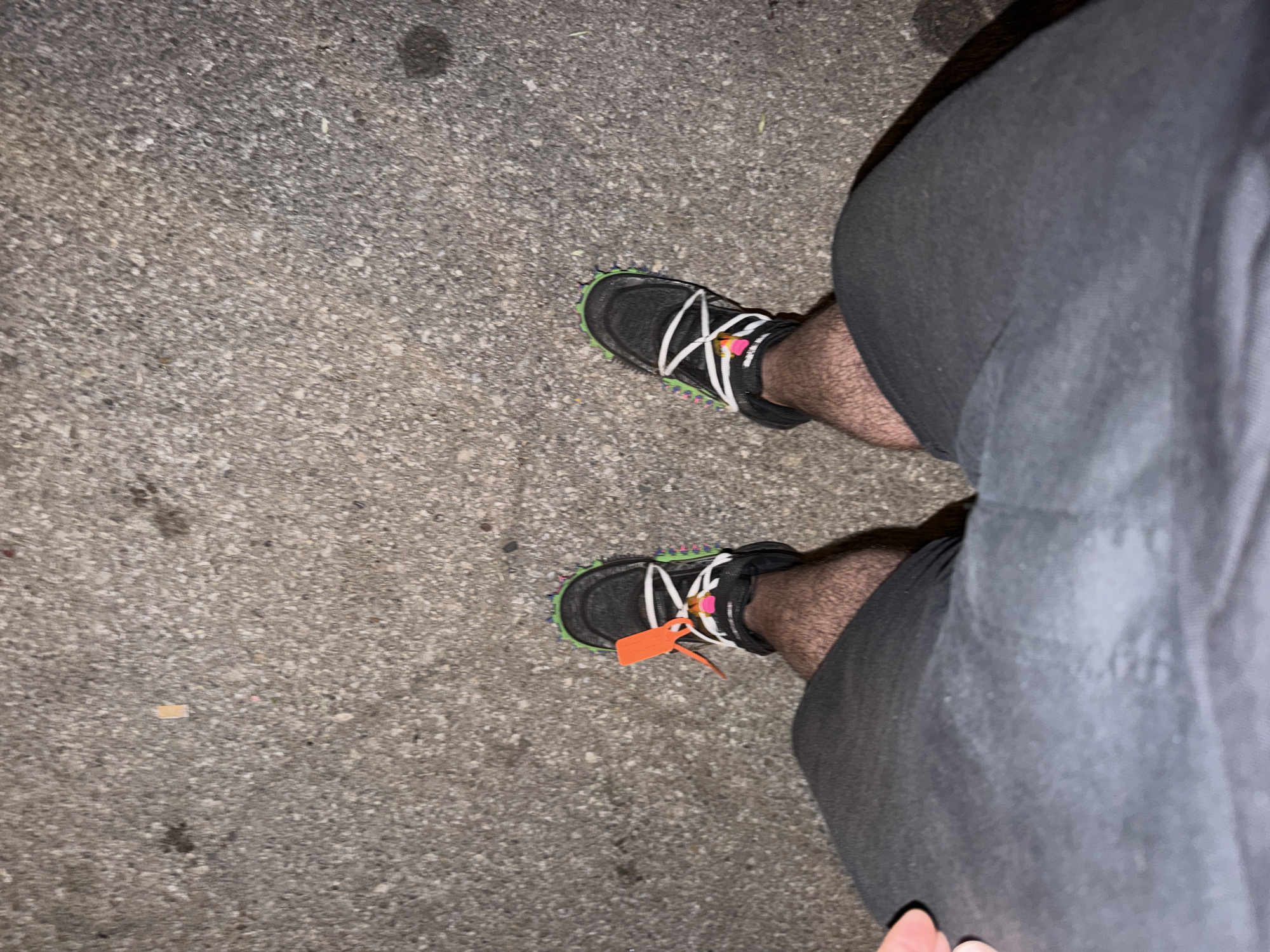 The image is taken from a high angle, showing a person standing on a concrete surface, possibly a sidewalk or road. The person is wearing dark gray shorts and black sneakers with white laces and a neon green sole. One of the sneakers has an orange tag attached. The concrete surface is gray and textured. There are no other identifiable people, objects, or details in the scene. The location cannot be determined to be Sofia, Bulgaria based on this image.