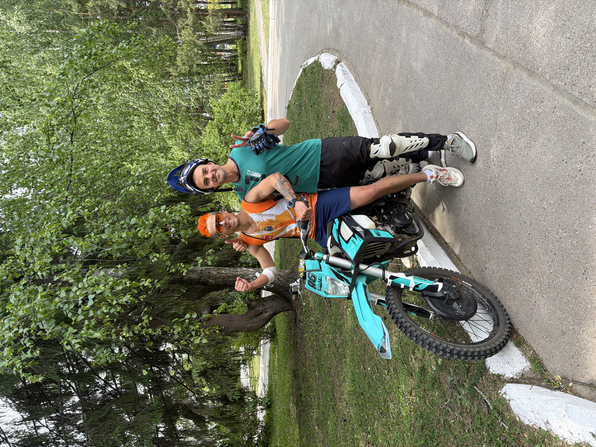 Two men are posing with a turquoise-colored Avantis dirt bike. One man, wearing an orange wig and sunglasses, is seated on the bike, giving a thumbs-up. The other man, wearing a helmet and knee pads, is leaning on the bike, giving a peace sign. They are on a path near the city of Chaikovsky, Russia. There is a curb, trees, and grass around them.
