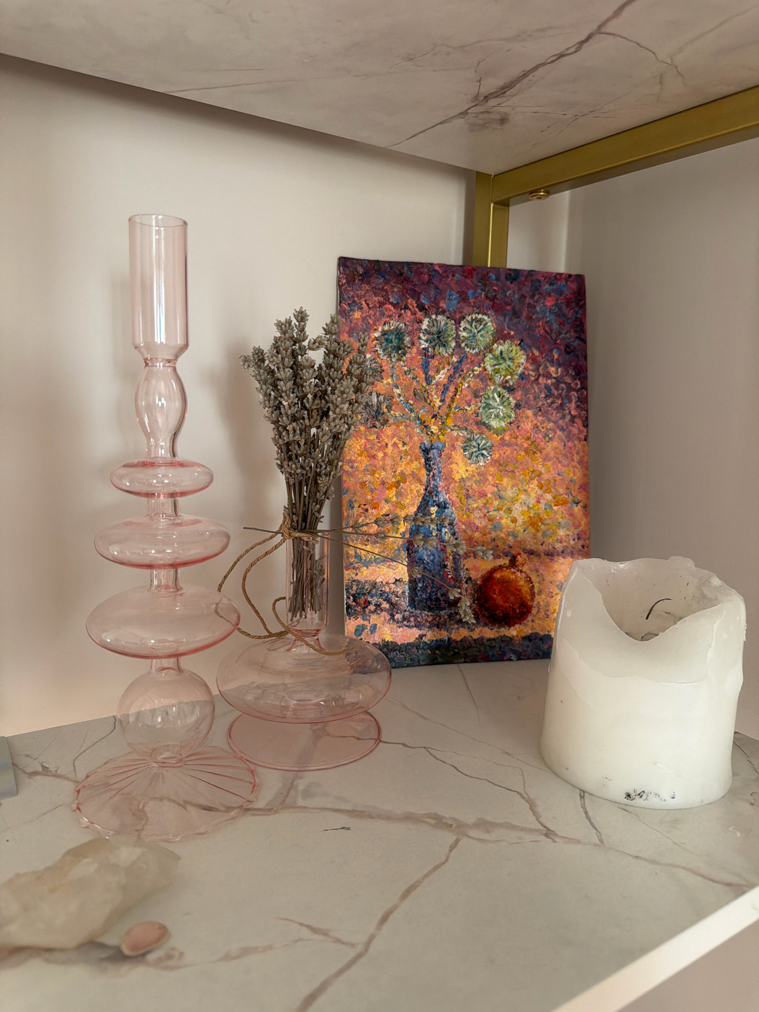This is a still life image set on a white marble surface, likely within a home. The scene features several decorative objects: a tall, tiered pink glass candlestick holder on the left, a small vase with dried lavender next to it, a colorful abstract painting of a vase of flowers and a round fruit on the right, and a large white pillar candle on the far right. The background appears to be a white wall with a hint of a marble-patterned shelf above. There are no people or animals visible. The lighting is soft and diffused. The location is described as Samara, Russia, but there are no visual cues in the image to confirm this specific location.