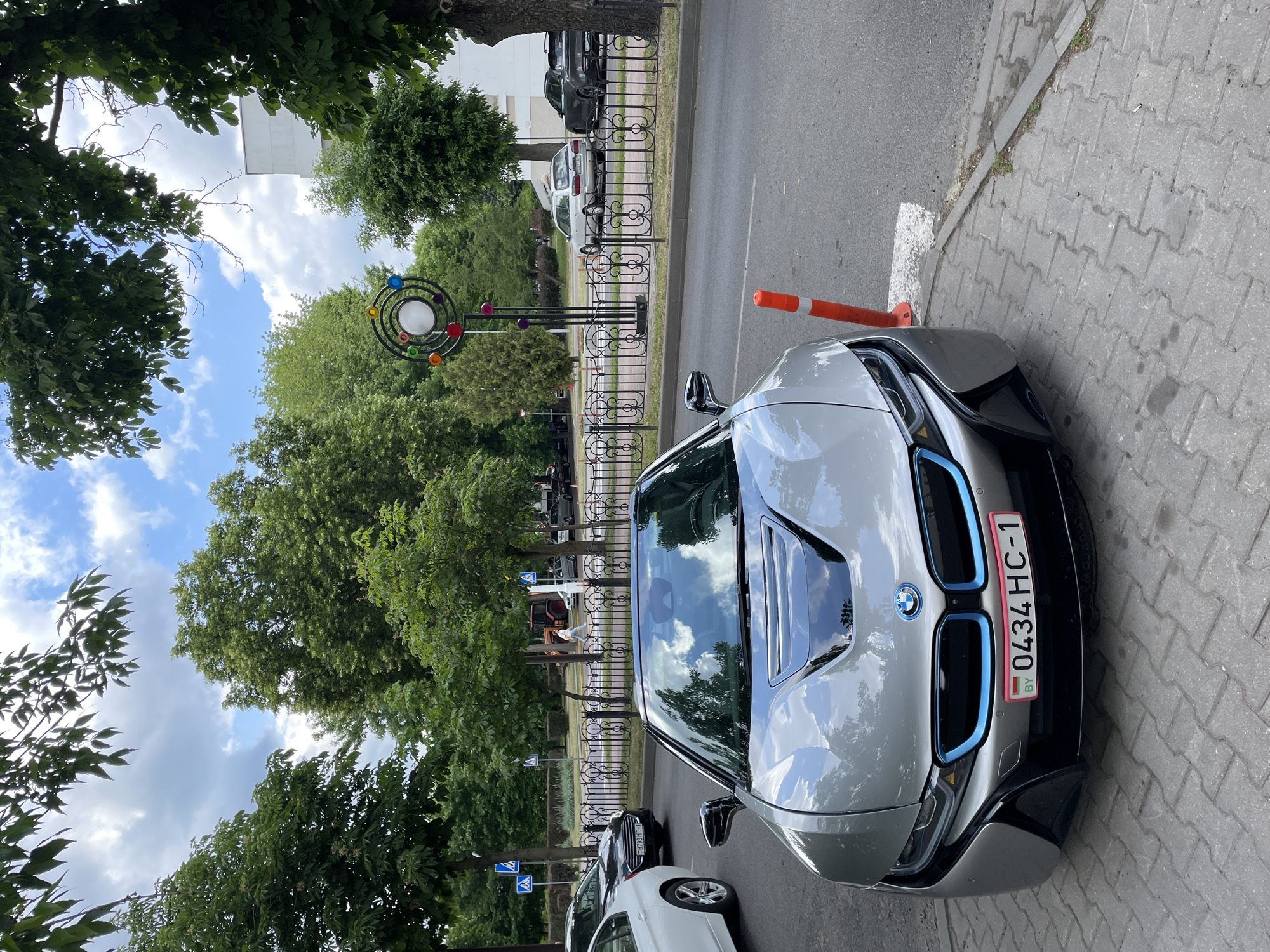 The image shows a silver BMW i8 parked on a brick sidewalk in Brest, Belarus. The car is partially blocking a crosswalk, with an orange traffic cone positioned next to its front bumper. The license plate reads "0434 HC-1" with the "BY" country code. Behind the car, a wrought-iron fence separates the sidewalk from the street. Trees line the street, and a decorative lamp post with colorful spheres is visible. Several other parked cars are visible in the background. The sky is blue with scattered clouds, reflected in the car's windshield.