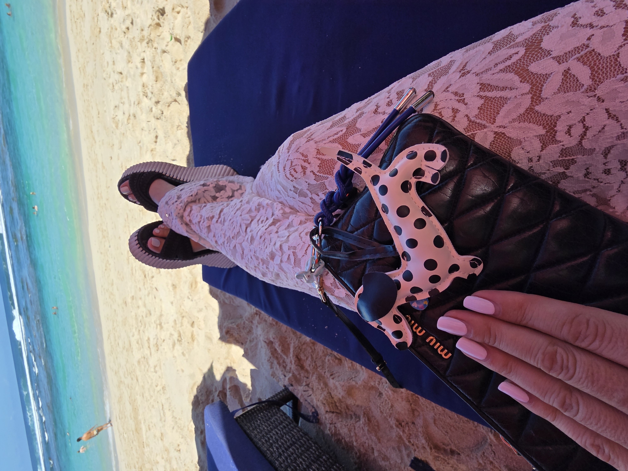 A woman sits on a lounge chair on a beach, presumably in Kuta Selatan, Indonesia, based on the turquoise water and sandy shoreline visible in the background. She wears a light-colored lace dress and black sandals with a thick, ridged sole. A black quilted purse, adorned with a white and black spotted dog keychain, rests on her lap. Her hand, sporting neatly manicured pink nails, partially covers the purse. The lounge chair has a dark blue cushion. The beach stretches out to the left, meeting the ocean.