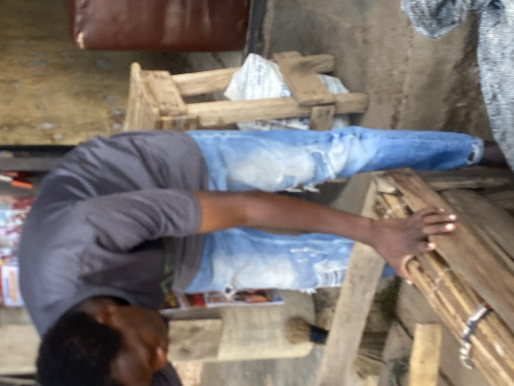 A man is bending down and handling a bundle of wooden sticks in what appears to be an outdoor market in Bukuru, Nigeria. He is wearing a grey t-shirt and light blue ripped jeans. To his right, a wooden stool with a bag on it is visible. The ground is dirt.