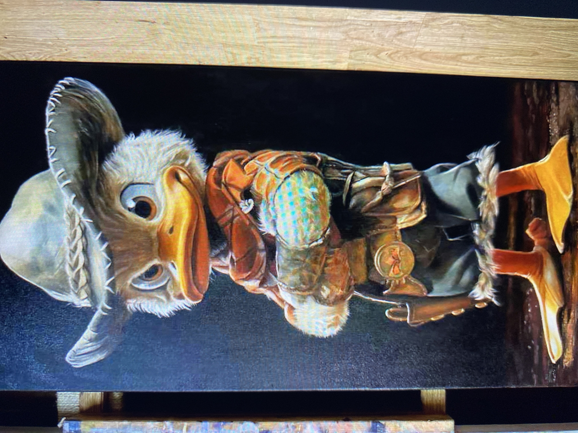 A full-body, vertical, painted portrait of a cartoon duck dressed as a cowboy is shown against a dark background. The duck wears a light-brown, wide-brimmed cowboy hat with a braided band. Its face is white and fluffy, with large, expressive blue eyes and an orange beak. The duck is adorned in a brown leather vest over a reddish-orange scarf, with lighter brown arm coverings and blue-green patterned gloves. A belt with a circular, gold buckle and a holstered pistol is visible around its waist. It wears dark green pants with fur trim and orange, webbed feet. The duck stands with its arms crossed and its expression is stern. The painting is framed by a light wooden structure on the right side of the image, and a portion of another framed artwork is visible on the left. The scene is likely indoors, given the controlled lighting and presentation of the artwork.