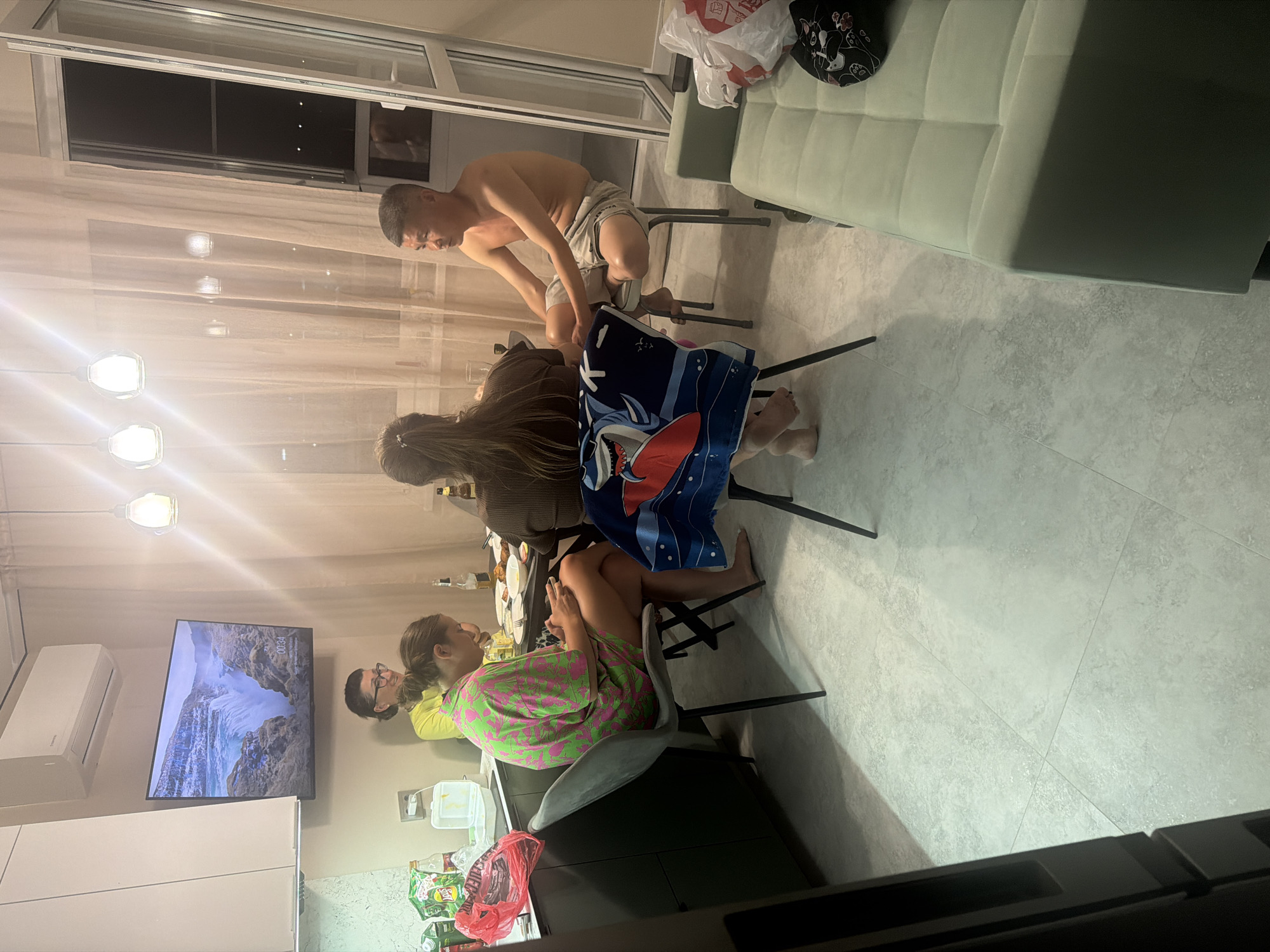 A group of four people are gathered around a table in what appears to be a small apartment kitchen or dining area. The room is illuminated by artificial light from overhead fixtures, and natural light from a balcony door that is partially open.
In the foreground, on the left, a woman wearing a bright green and pink patterned dress is seated at a chair, looking down at her phone. She is barefoot. To her right, another woman with long brown hair pulled back in a ponytail is also seated, facing away from the camera and towards the table.
At the table, a man with short dark hair is seated, shirtless, wearing light-colored shorts. He is looking towards the left side of the frame. Another person, partially obscured by the man, is also seated at the table.
On the table, there are plates with food, bottles, and glasses, suggesting a meal or gathering is in progress. To the left of the table, on a counter, are various items including bags, a white container, and bottles.
Above the counter, a flat-screen television is mounted on the wall, displaying a scenic image of a waterfall. An air conditioning unit is also visible on the wall above the television.
To the right of the scene, a couch with a light green upholstery is visible. A black cap with a white cat design is placed on the couch. A white plastic bag with red lettering is also on the floor near the couch.
The floor is tiled with a light gray pattern. The walls appear to be painted a light color, and sheer curtains hang behind the table area. The balcony door shows a view of the night sky.
The image does not provide any specific visual cues to definitively place the location within the city of Novorossiysk, Russia.