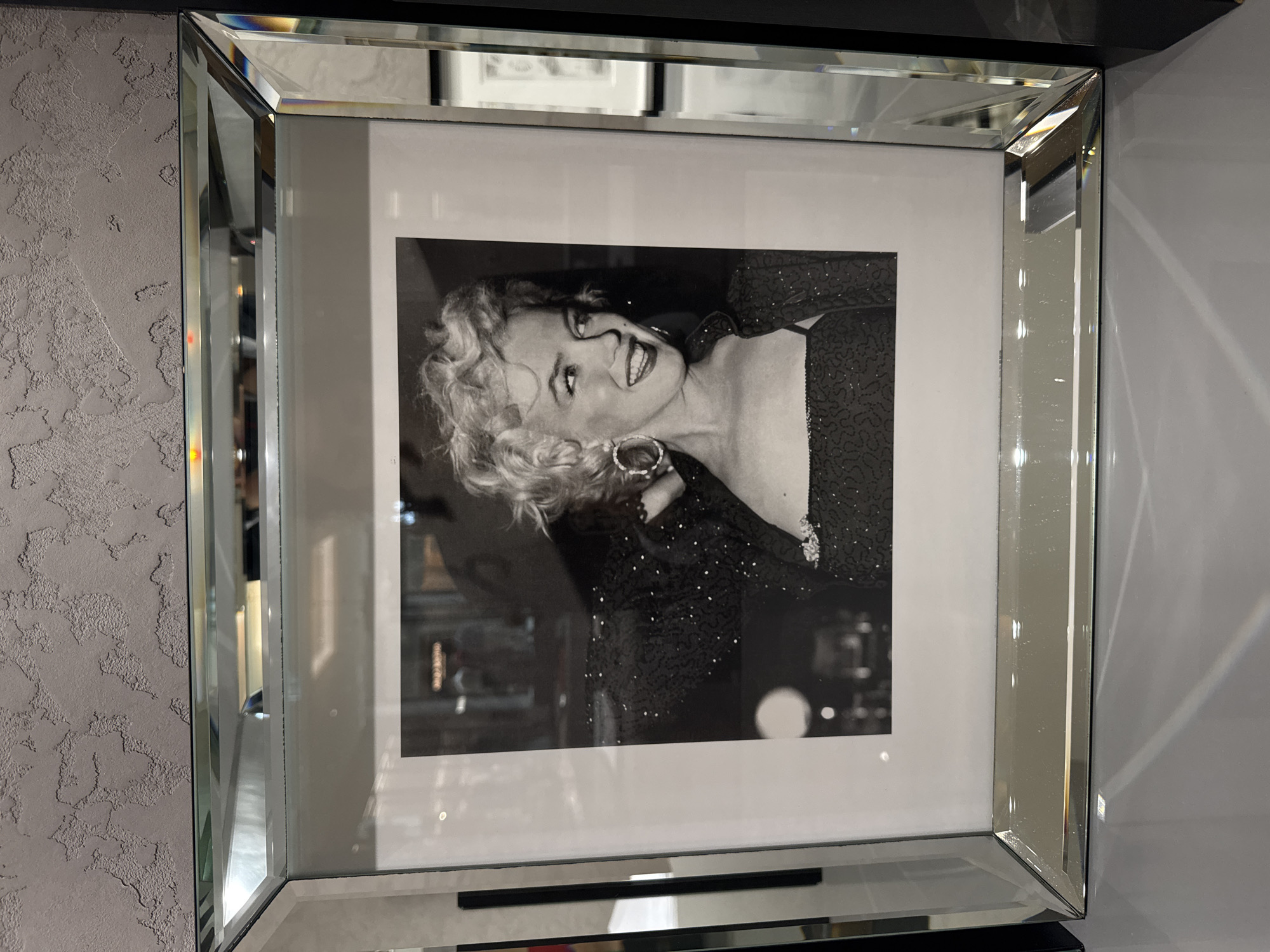 A black-and-white photograph of a smiling woman with short, curly hair wearing a sparkly dress and large earrings is framed in a mirrored frame with beveled edges. The photograph is hung on a wall; to the left of the frame, the wall is textured and gray, while the wall to the right is smooth and white. Above the frame, another picture is partially visible. The location is Dubai, United Arab Emirates.