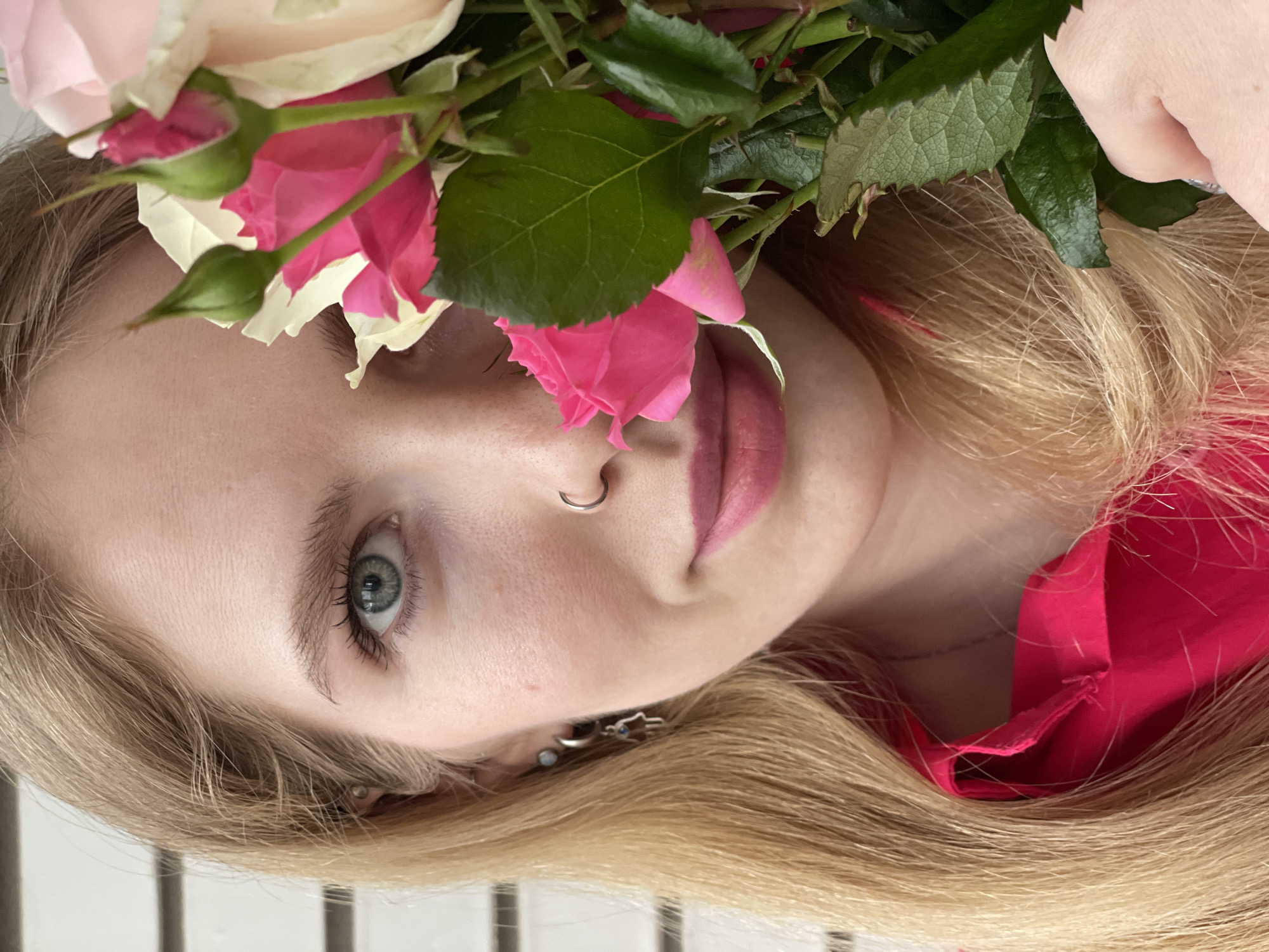 A close-up, angled portrait features a fair-skinned woman partially obscured by pink and white roses with green foliage. The woman has blond hair, a nose ring, and noticeable earrings. She wears a red top. The background consists of white horizontal lines. There is no indication this photo was taken in Izhevsk, Russia.