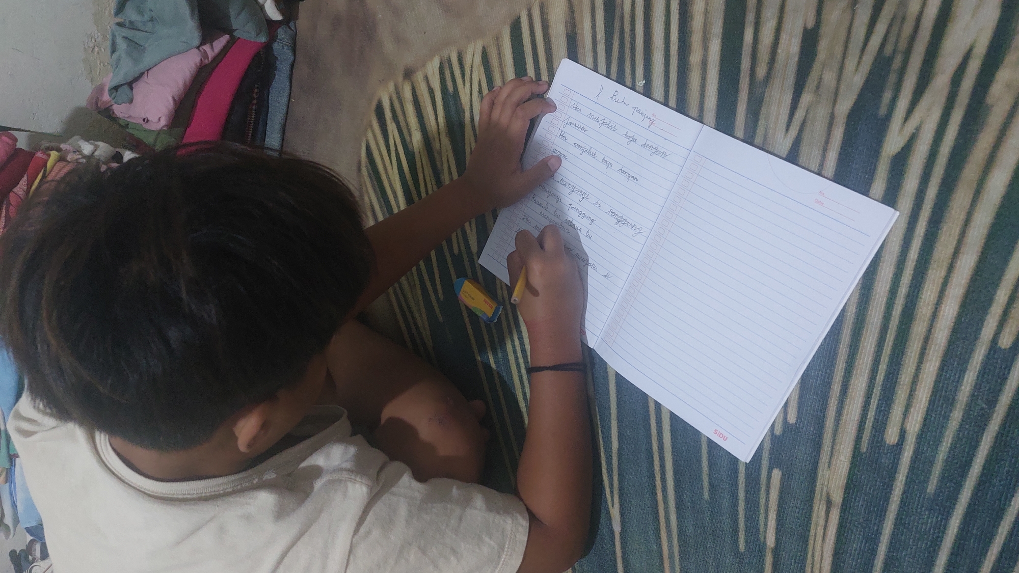 A young person is seen from a high angle, lying on their stomach on a patterned mat and writing in a lined notebook with a yellow pen. The person is wearing a light-colored t-shirt and has dark hair. To the right of the notebook, a blue and yellow eraser is visible. In the background, a pile of colorful folded clothes rests against a wall, suggesting a domestic setting, likely in Jatirogo, Indonesia, given the context. The lighting is dim, casting shadows and obscuring some details.